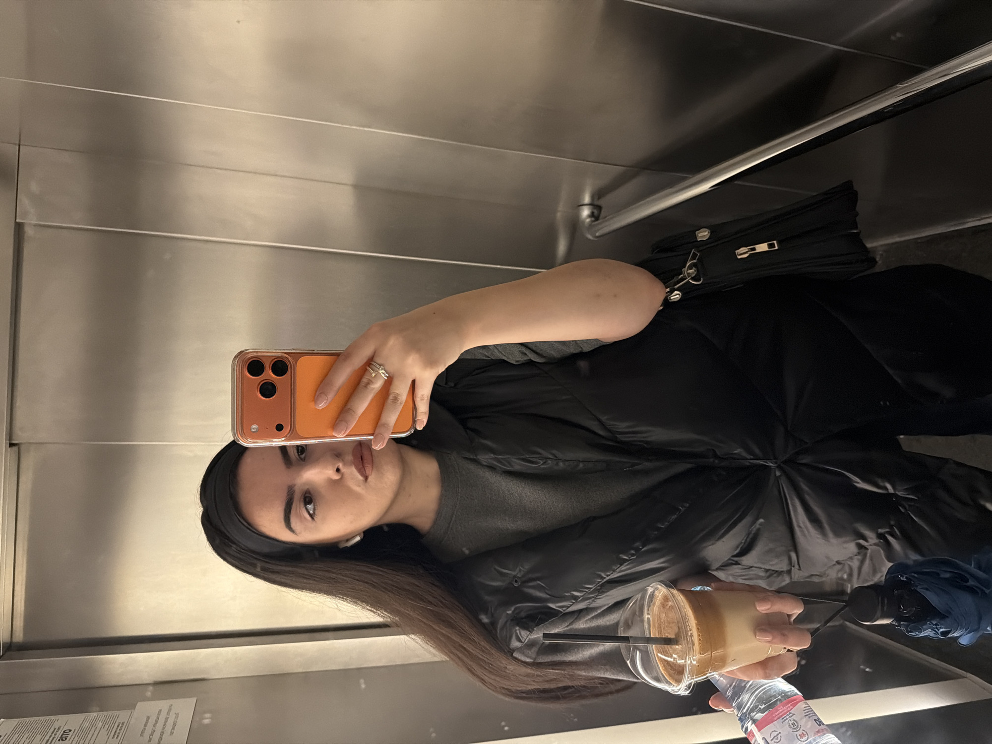 The image displays a mirror selfie of a young woman inside what appears to be an elevator or a small, enclosed space with reflective metallic walls, indicative of a location in Sofia, Bulgaria. The woman, with long dark hair and a dark headband, is wearing a dark puffer jacket over a gray long-sleeved top. She holds an orange-cased smartphone (likely an iPhone given the camera module design) up to take the picture, capturing her neutral expression. Her nails are painted a light color, and she wears a ring on her left hand.
In her right hand, she holds a clear plastic cup containing a layered iced coffee or similar beverage with a black straw. Below the drink, she also holds a clear plastic bottle of water. A black shoulder bag with silver hardware is visible next to her. The metallic walls create reflections, and a horizontal handrail is present on the right side of the frame.
Visible text on the water bottle includes "AQUAFINA" and "DEVIN," along with Cyrillic script such as "24 ЧАСА" (24 hours) and "0.5л" (0.5 liters). On a notice or sticker on the bottom left wall, blurred Cyrillic text is visible, with the word "ПОСЛЕДНИ" (last) faintly distinguishable. The image captures a casual moment, likely during transit.
