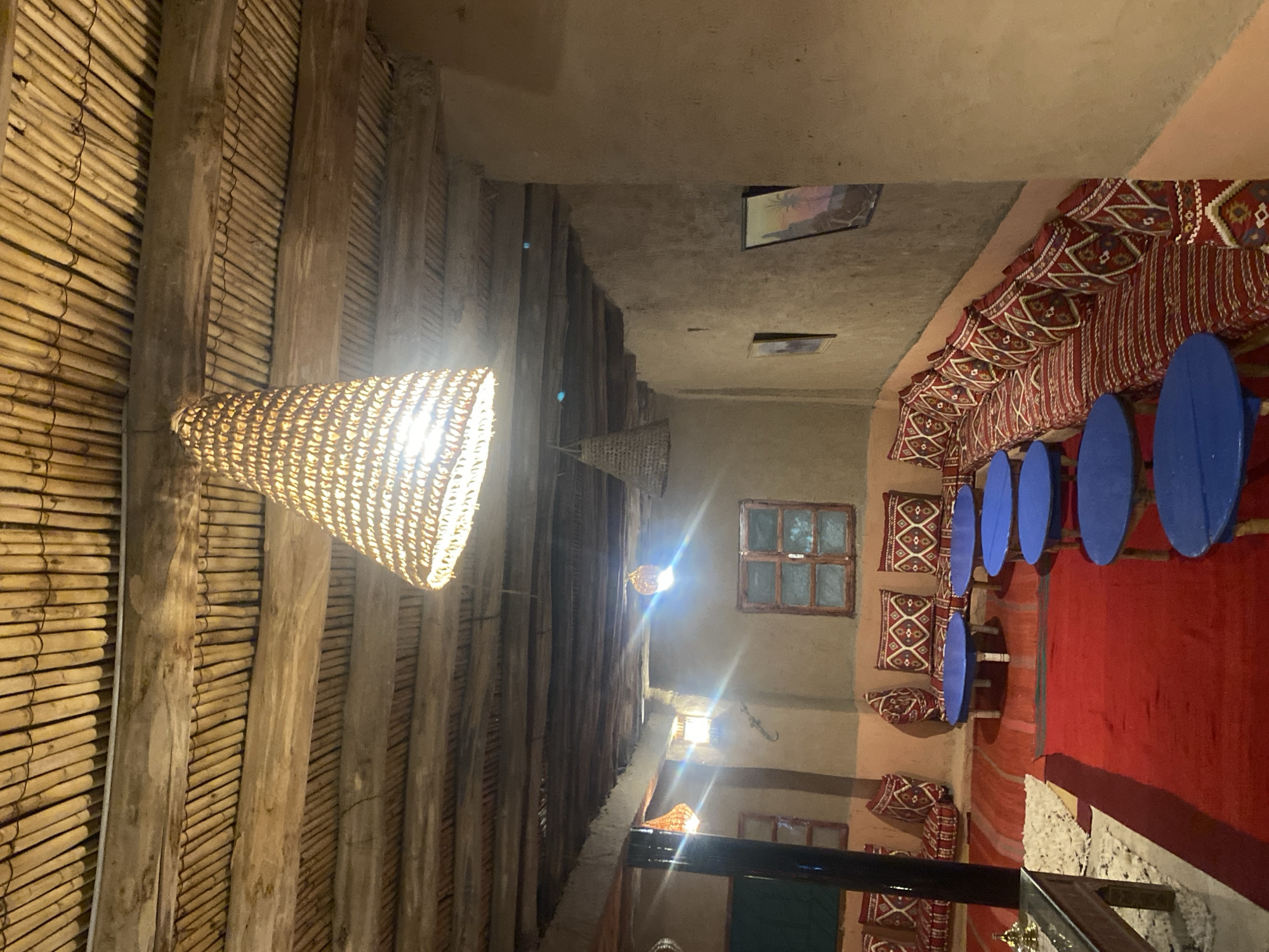 This is an interior scene of a traditional Moroccan tent or riad in Agafay, Morocco. The ceiling is constructed from exposed wooden beams and bamboo poles, with a large, woven conical lamp hanging from the center. The walls are made of natural clay or mud plaster, giving the space a rustic feel. Along the walls are built-in benches covered with red and white patterned cushions, creating a comfortable seating area. In front of the benches are low, round blue tables. Several other woven lamps are visible, as well as framed artwork on the walls. Red carpets cover the floor, adding warmth and color to the space. The overall ambiance is one of a cozy, authentic Moroccan dwelling, likely a restaurant or guesthouse.