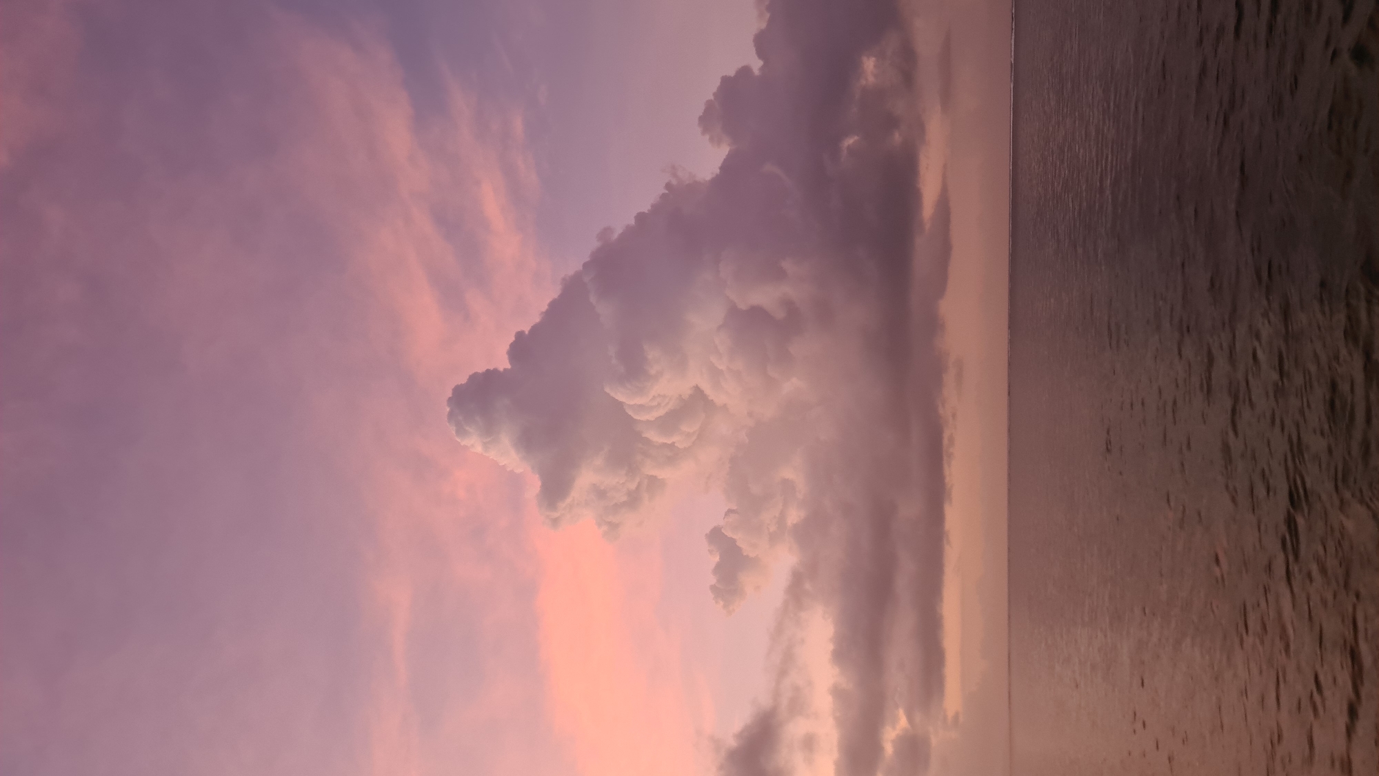 This is a vertical, low-angle photograph of a sunset over the ocean, taken from Denpasar, Indonesia. The image is dominated by a vast expanse of sky, painted with the soft hues of twilight. Wispy clouds, tinged with shades of pink and purple, stretch across the upper portion of the frame, their ethereal forms contrasting with a larger, more dramatic cumulonimbus cloud formation that stands out in the center. This massive cloud, with its textured, cauliflower-like appearance, is illuminated by the fading sunlight, casting shadows and giving it a sense of depth and volume.
Below the sky, the ocean stretches horizontally across the lower half of the image, appearing calm and reflective. The water's surface is a deep, muted purple, mirroring the colors of the sky and subtly disturbed by small ripples. The horizon line, where the sky meets the sea, is a thin, indistinct band, suggesting a wide, open vista. The overall mood of the image is serene and peaceful, evoking the tranquil beauty of a sunset. The time of day is clearly sunset, given the vibrant colors in the sky and the soft lighting. There is no visible text in the image.