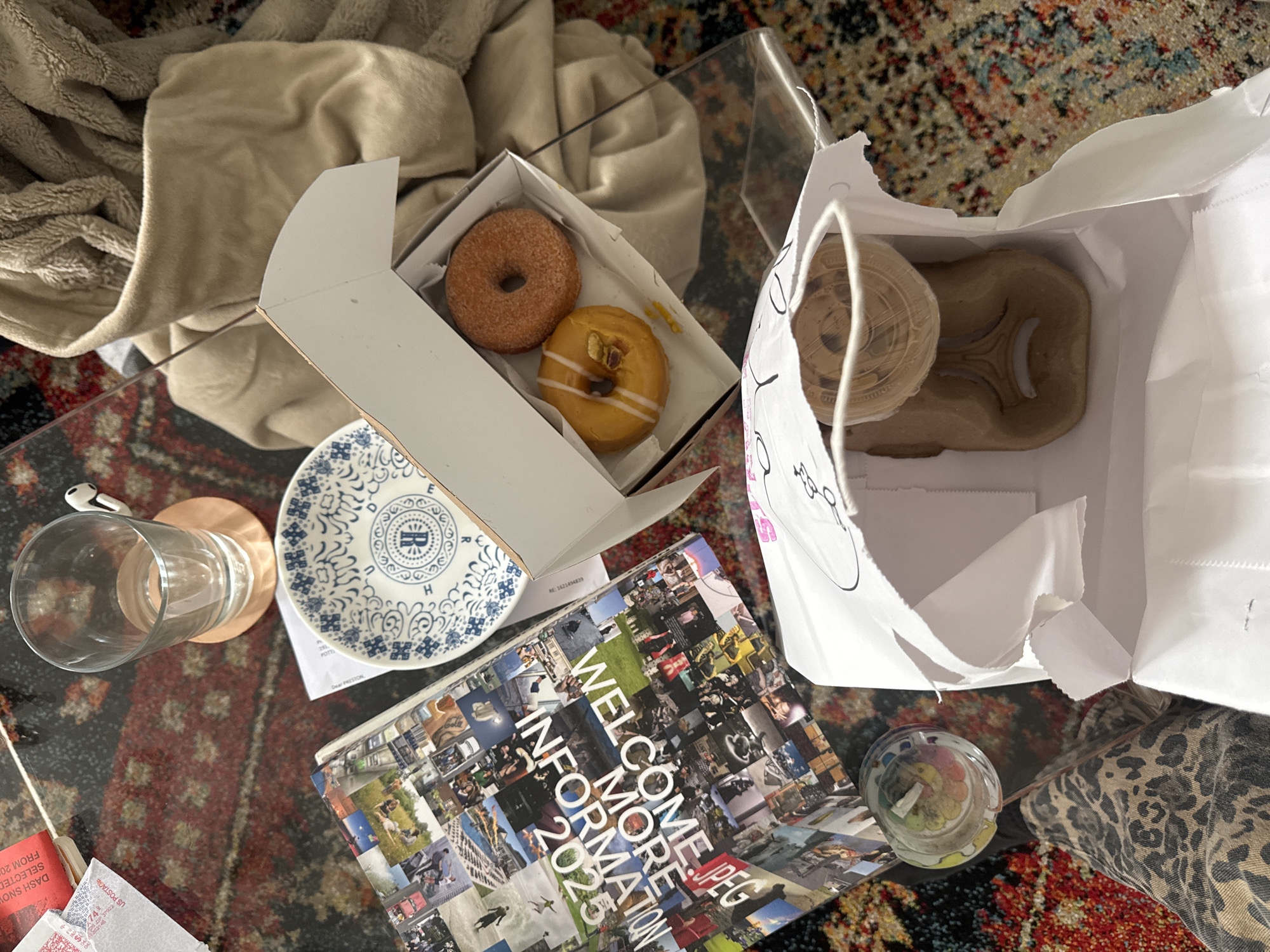 The image captures a casual, overhead view of items resting on a clear glass coffee table, set over a vibrant, patterned rug with shades of red, blue, and black. In the upper left, a soft, beige blanket is draped over what appears to be a couch or chair, hinting at a comfortable indoor setting.
The focal point is an assortment of food and drink. Two donuts are presented in an open white box: one is coated in cinnamon sugar, and the other features a yellow glaze, drizzled with white icing, and topped with what looks like chopped pistachios. Beside the donut box, a white paper bag contains a takeaway coffee cup with a clear plastic lid and a brown cup carrier, suggesting a recent purchase.
On the table, an empty clear glass sits on a light wooden coaster, adjacent to a small, decorative blue and white plate with a distinctive 'B' initial and ornate patterns. Below these, a magazine or book titled "WELCOME MORE INFORMATION 2025" with "JPEG" also visible, displays a collage of numerous small photographs on its cover. Nearby, several pieces of mail are scattered, including an envelope marked "US POSTAGE". A small, clear, spherical object with colorful contents, possibly a decorative item or candle, is positioned to the right of the book.
Visible text includes "WELCOME MORE INFORMATION 2025" and "JPEG" on the book, "US POSTAGE" and "DASH SNOW SELECTED FROM 20" on mail, and "RE: 1621494839" on a piece of paper. The plate shows partially visible letters like "R", "B", "H", and "DER". The scene suggests a relaxed daytime moment, possibly a breakfast or snack, in a home in New York, United States of America.