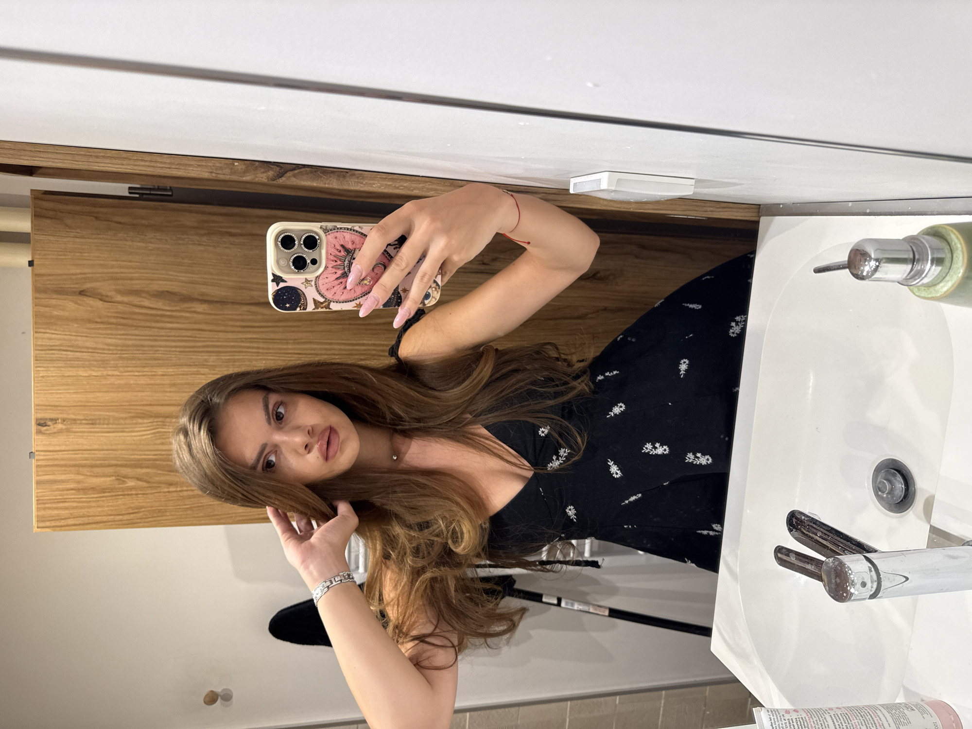 A young woman with long, wavy brown hair is taking a mirror selfie in a bathroom. She is wearing a black dress with white floral embroidery and a silver bracelet. She is touching her hair with her left hand and holding a pink and gold phone case in her right hand. In the background, there is a wooden cabinet, a white sink with a chrome faucet, and a white wall. The scene is illuminated by artificial light.