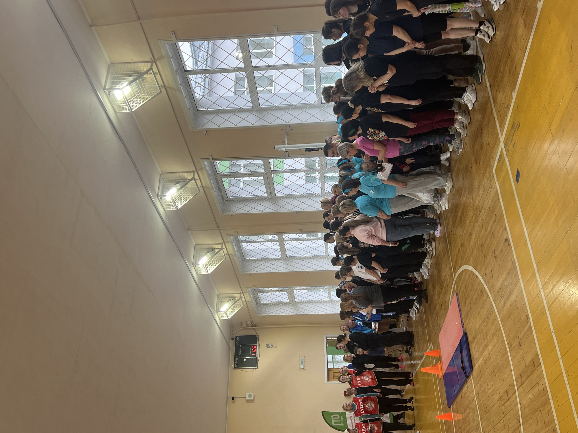 A wide, slightly tilted, indoor shot shows a large group of people standing in rows in what appears to be a gymnasium or indoor sports hall in Chaikovsky, Russia.
The space is well-lit with ceiling lights housed in wire cages. Tall windows with lattice patterns are visible along one side of the hall. The floor is made of wooden planks marked with white lines, suggestive of a basketball or volleyball court.
The group of people is diverse in age, with a mix of genders. They are arranged in rows. Some people are wearing blue shirts, while others wear black or other colors. At the front of the group are individuals wearing red vests with the word "СУДЬЯ" printed on them, which likely means "JUDGE" in Russian. A green banner with Russian text is also visible at the front.
At the far end of the gymnasium, a digital scoreboard displays numbers and words in Russian. Closer to the foreground, there is a gymnastic mat and several orange cones.