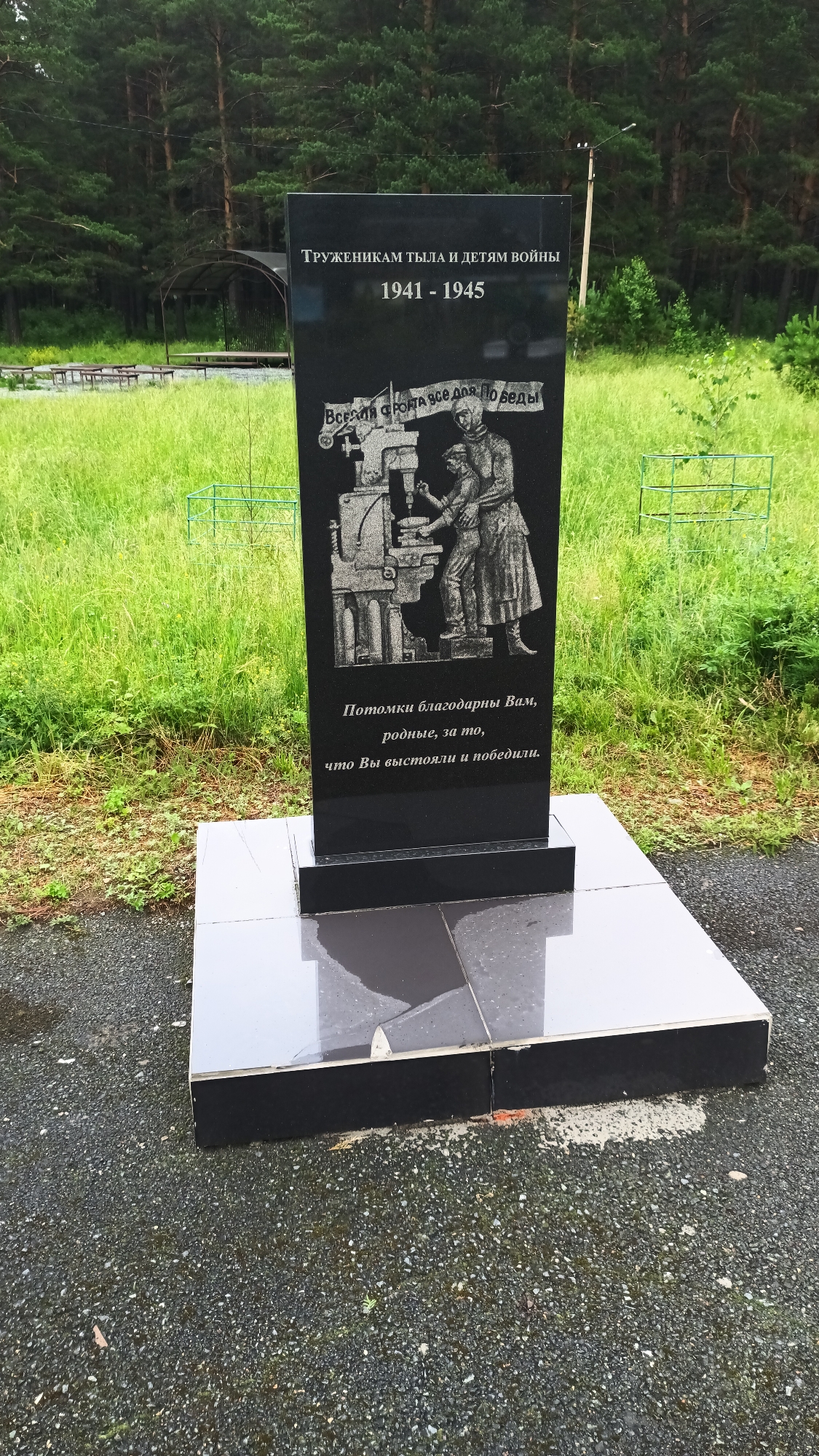 A black granite monument stands in a grassy area with trees in the background. The monument is rectangular and stands on a tiered base. The inscription at the top reads "ТРУЖЕНИКАМ ТЫЛА И ДЕТЯМ ВОЙНЫ" (To the workers of the rear and children of war) followed by the years "1941 - 1945". Below this is an etched image of two figures working at a machine. The inscription at the bottom reads "Потомки благодарны Вам, родные, за то, что Вы выстояли и победили." (Descendants are grateful to you, dear ones, for holding out and winning.). The area around the monument is covered in dark grey gravel, and the grass is green and somewhat overgrown. Faintly visible in the background is a dark green metal structure with tables and benches. This monument is located in Abaza, Russia, and it commemorates those who contributed to the war effort during World War II.