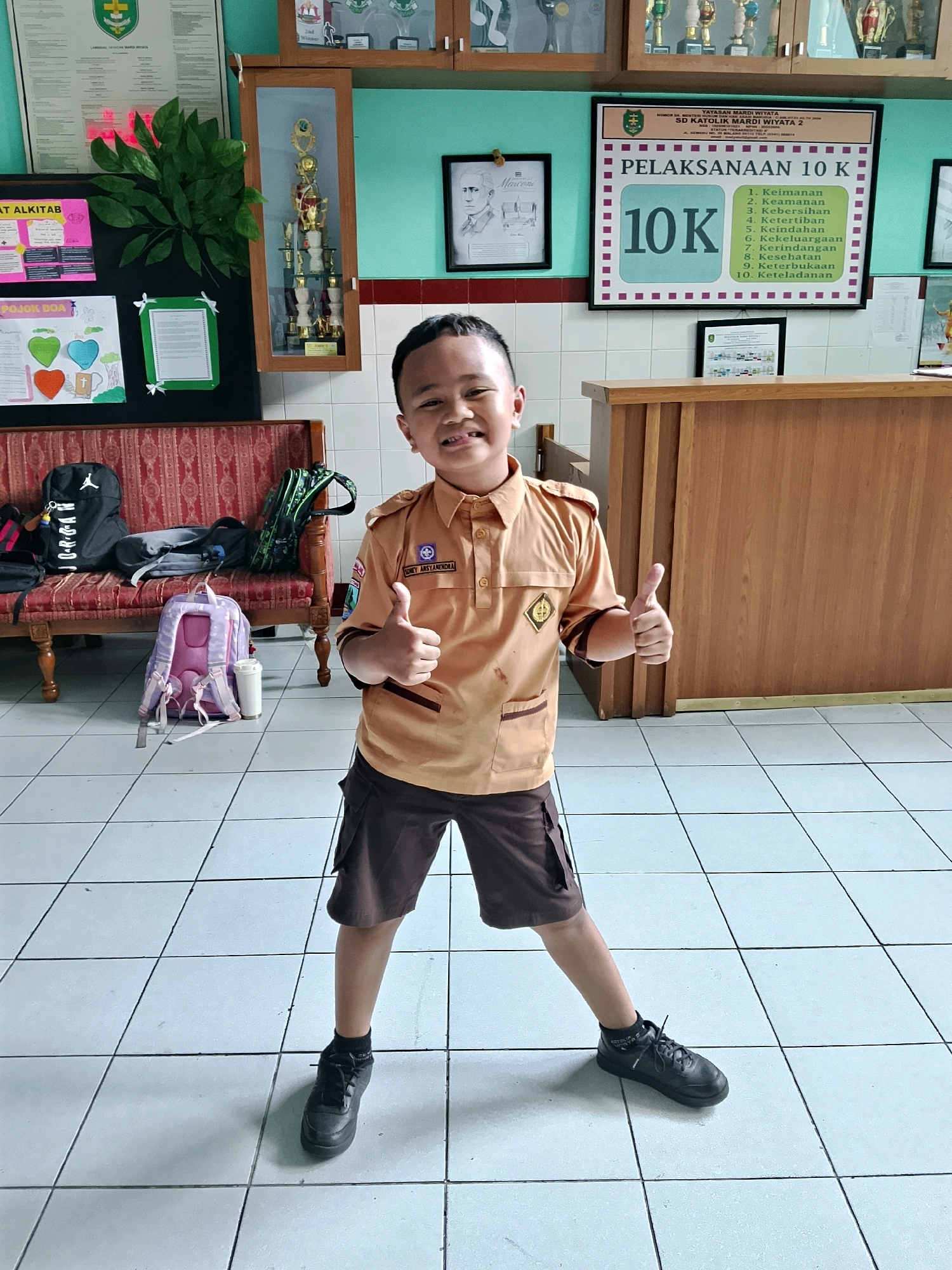 A young Indonesian boy, identified by the name tag on his uniform as "DONEY ARSYANDRA," stands confidently in a school hallway, giving a double thumbs-up. He is wearing a tan-colored uniform shirt with a school emblem on the left chest and a dark brown skirt with pockets. He has dark hair and is smiling broadly at the camera.
The hallway features a light blue painted wall, a wooden reception desk with vertical paneling, and a tiled floor. Several objects are visible in the background, including backpacks on a patterned red bench, trophies displayed in a glass-fronted cabinet, framed pictures, and educational posters. One poster, prominently displayed, reads "PELAKSANAAN 10 K" which translates to "10K Implementation" and lists ten points related to school conduct or values.
The environment suggests a school setting, likely in Malang, Indonesia, given the context of Indonesian school uniforms and the name "SD KATOLIK MARDI WIYATA 2" visible on one of the posters. The lighting is bright, indicating indoor lighting. The overall atmosphere is cheerful and positive.