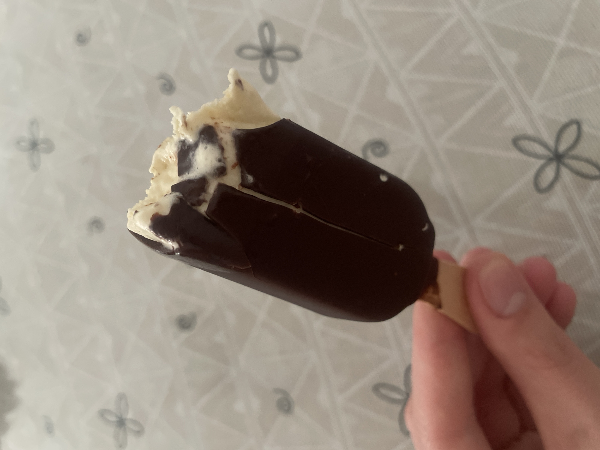 A hand is holding a partially eaten chocolate-covered ice cream bar. The ice cream is vanilla flavored with chocolate chunks. The background is a white tablecloth with a subtle grey floral pattern. This image was captured indoors, likely in a home environment, in Atarfe, Spain.