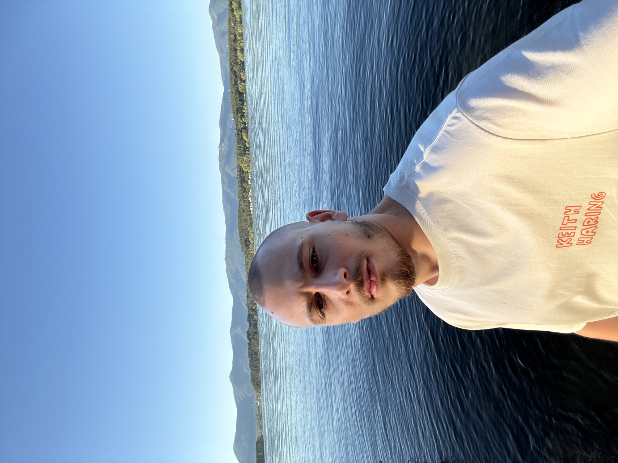 A man with a shaved head and a goatee is taking a selfie by the water. He is wearing a white t-shirt with red lettering that reads "KEITH HARING". The man is positioned in the foreground, with the water and distant mountains in the background. The water is a deep blue with ripples, reflecting the clear sky. The mountains are green and covered in trees, with some hints of structures on them. The sky is a bright, clear blue. The overall scene suggests an outdoor recreational setting, possibly a lake or reservoir. The location is identified as Koprinka, Bulgaria.