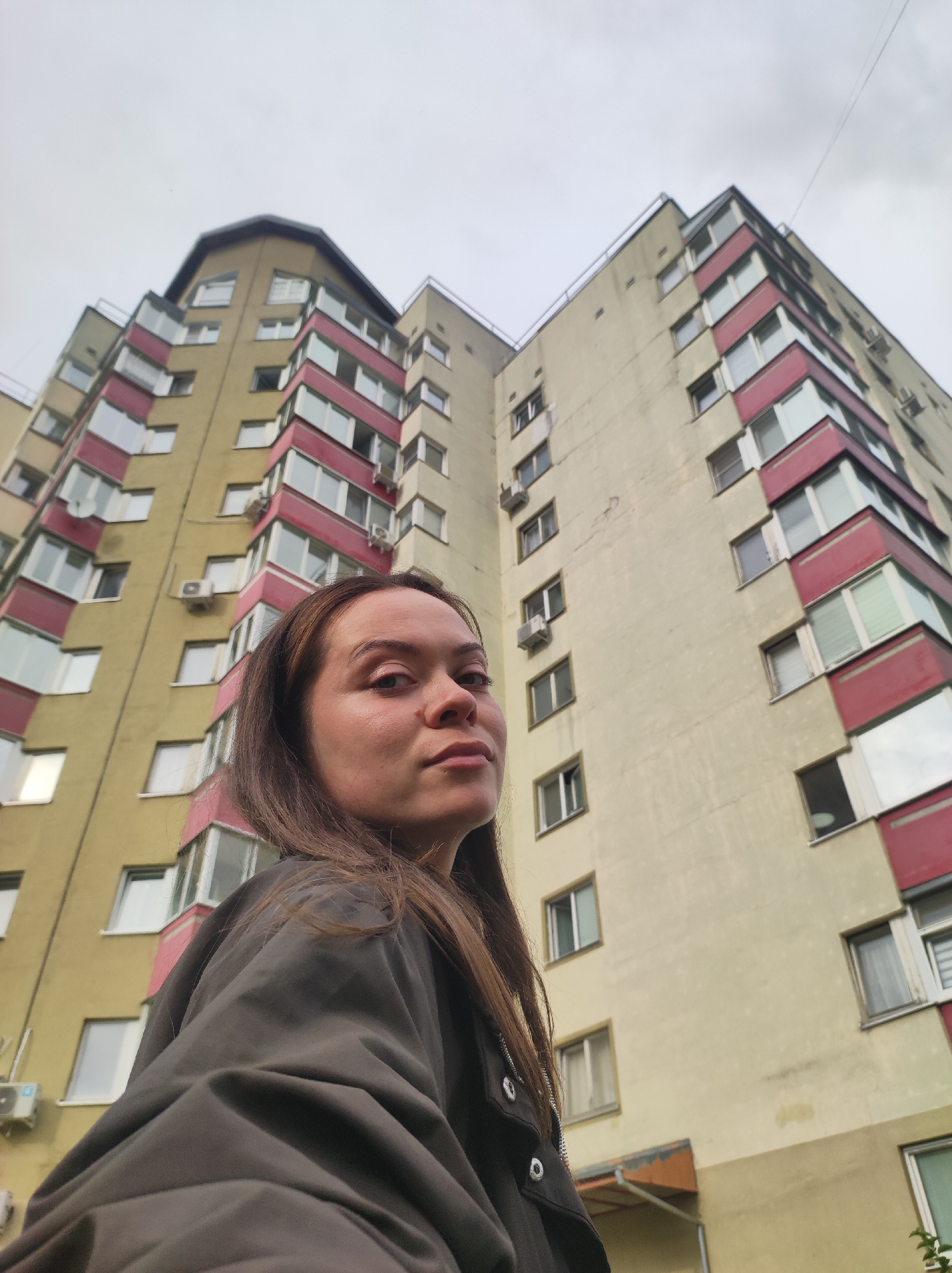 An upward-angle, medium shot captures a young woman in the foreground, looking directly at the camera. Her long, brown hair falls over her shoulders and is slightly windswept. She is wearing a dark, collared jacket. Behind her, a multi-story apartment building dominates the background. The building is painted a pale yellow with maroon accents around the balconies and windows. The sky above is overcast and grey. The scene appears to be in an urban environment, consistent with an apartment complex in Chaikovsky, Russia.