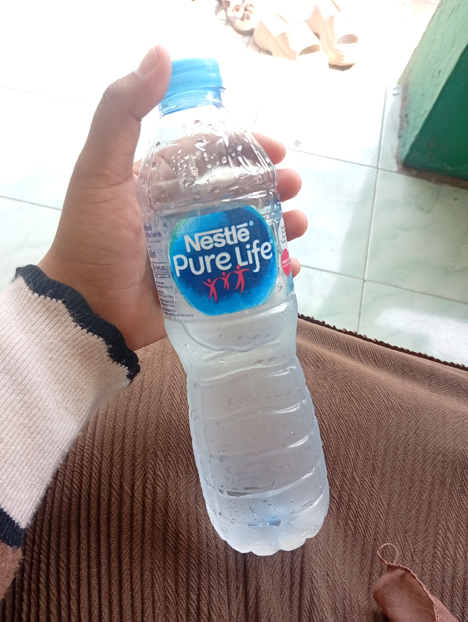 A person's hand, with a thumb and index finger visible, holds a partially full, condensation-covered plastic bottle of Nestlé Pure Life water. The bottle has a blue cap and a blue and white label featuring the Nestlé logo and the words "Pure Life" in white text, with stylized red figures below. The person is wearing a beige garment with a black and navy blue striped cuff on their left arm. The bottle is positioned over a textured brown fabric. In the background, a light-colored tiled floor is visible, along with what appears to be a green wall or structure. Additionally, a pair of light-colored sandals or flip-flops can be seen in the upper background. The scene is likely indoors or in a sheltered outdoor area with a tiled floor, common in residential or public spaces in cities like Sarang, Indonesia.