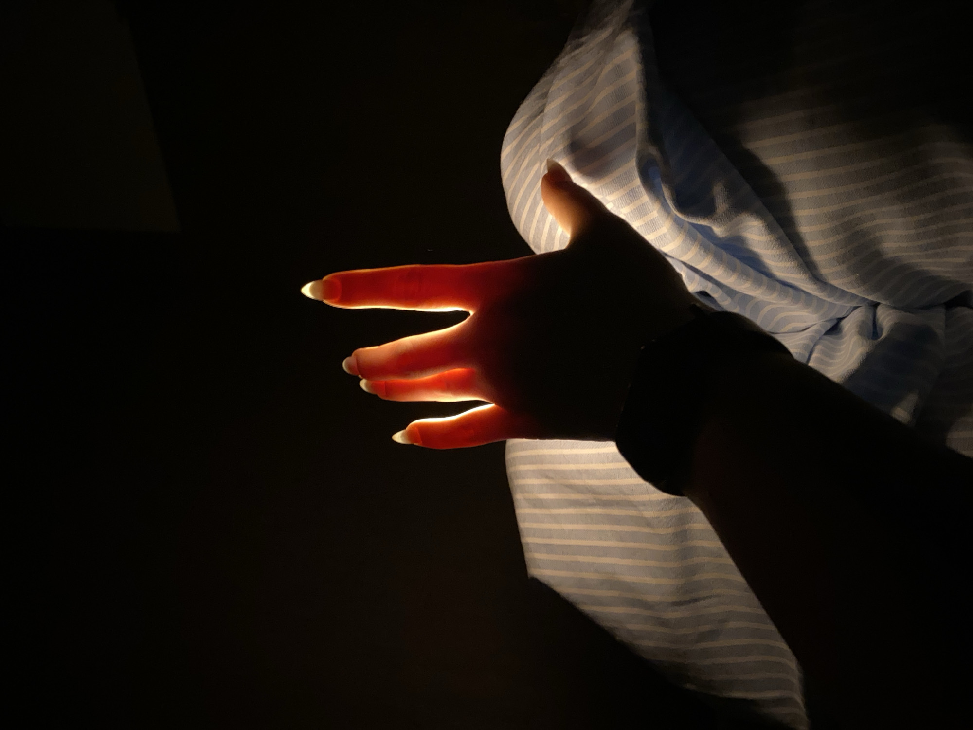 A hand is illuminated from behind, revealing its structure and long nails. The hand is positioned in front of a blue and white striped cloth. The background is dark. There is no information in the image to identify the location as Chaikovsky, Russia.