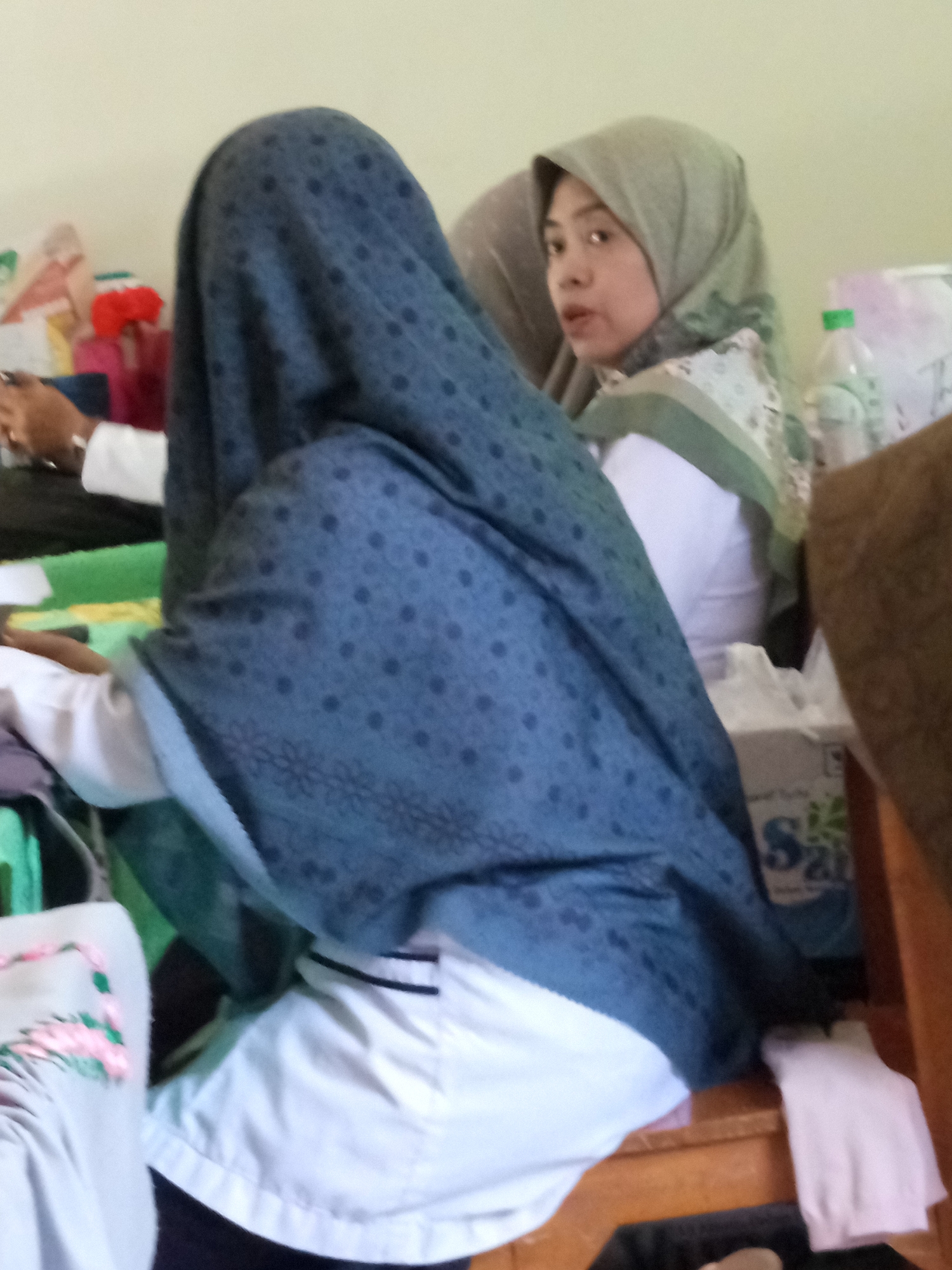 Two individuals are present in the image, both wearing hijabs. One individual, in the foreground, is largely obscured by a patterned blue hijab and white clothing. The other individual, in the background, is wearing a light green hijab and white clothing, and is looking towards the camera. They are seated on a wooden bench or chair in what appears to be an indoor setting. Various items are visible in the background, including what looks like a box with branding, plastic bottles, and fabric items. The overall impression is of a casual gathering or indoor activity. The image does not provide specific visual cues to determine the exact location within Lumutan, Indonesia, but it depicts a domestic or communal indoor space.