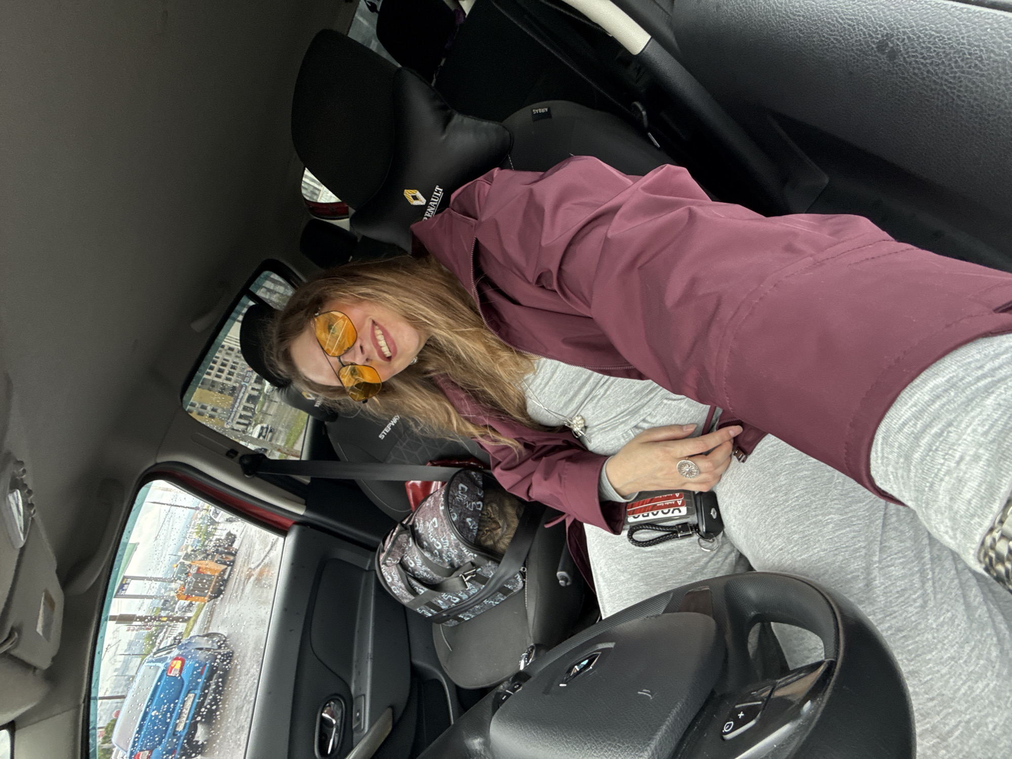 A woman is sitting in the driver's seat of a car, looking towards the camera. She is wearing sunglasses and a burgundy jacket. Inside the car, there is a pet carrier in the passenger seat. Outside the car, it appears to be raining. Through the window, buildings are visible. Based on the image and context, the location appears to be Izhevsk, Russia. A blue car is seen behind the subject's car. An orange construction vehicle is also visible outside the car.