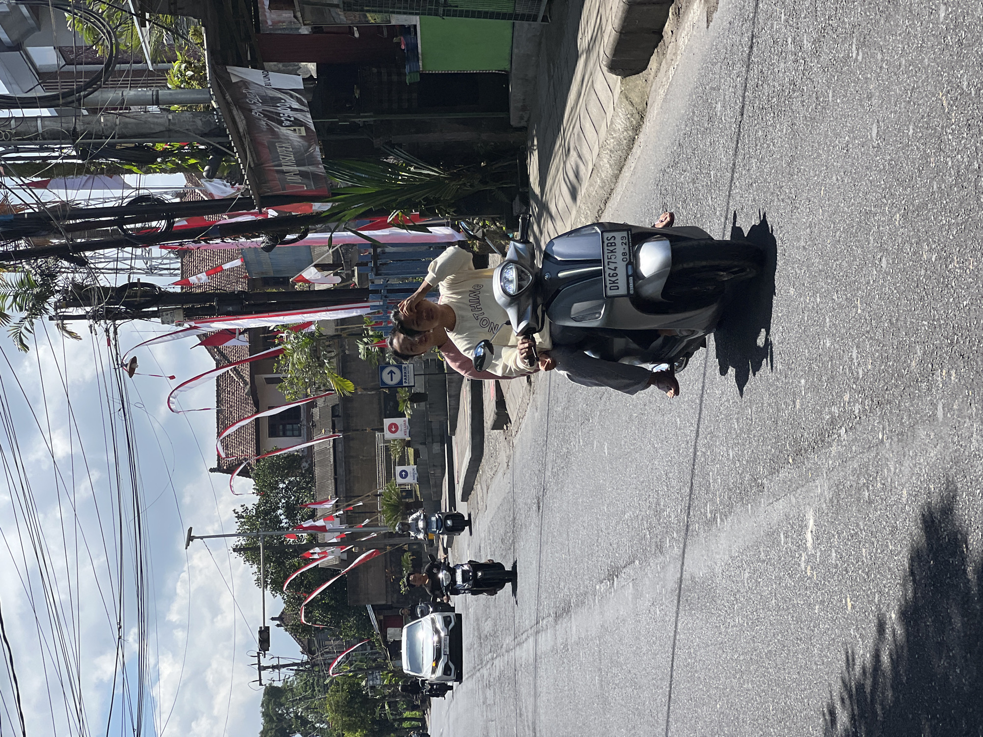 In Payangan, Indonesia, two individuals ride a silver scooter down an asphalt road. The scooter has a license plate that reads "DK6475KBS 08-29". The person in the front is wearing a white sweatshirt with "NOTHING" written on it and gray pants. The person in the back is wearing a pink shirt and gray pants. They are both looking forward, and the person in the front is shielding their eyes from the sun. Red and white Indonesian flags are strung along the roadside, attached to poles and wires. Buildings with red-tiled roofs are visible in the background. Other vehicles, including a white car and several other scooters, are on the road. The sky is partly cloudy.