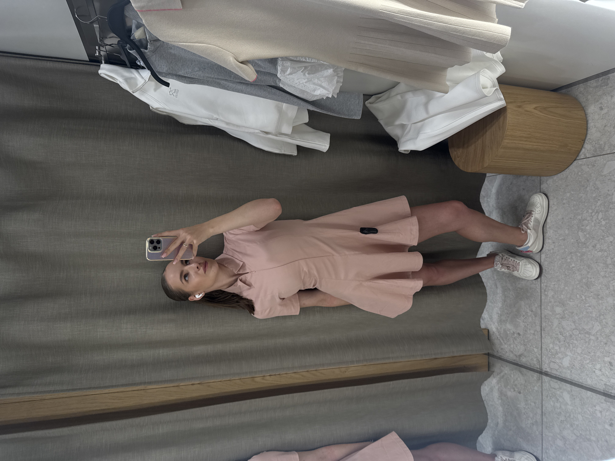 The image shows a young woman standing in a fitting room, taking a selfie with her phone. She is wearing a pink short-sleeved dress and white sneakers. Her hair is pulled back, and she is wearing wireless earbuds. The fitting room has grey curtains, a wooden stool, and tiled flooring. Behind her, clothes are hanging on a rack, including a grey zip-up jacket and white garments. The lighting is bright, suggesting it might be daytime. The woman appears to be looking at her phone, possibly checking her appearance or the photo. The overall scene suggests a shopping or trying on clothes scenario. There is a brand logo visible on the dress, but the text is too small to read.