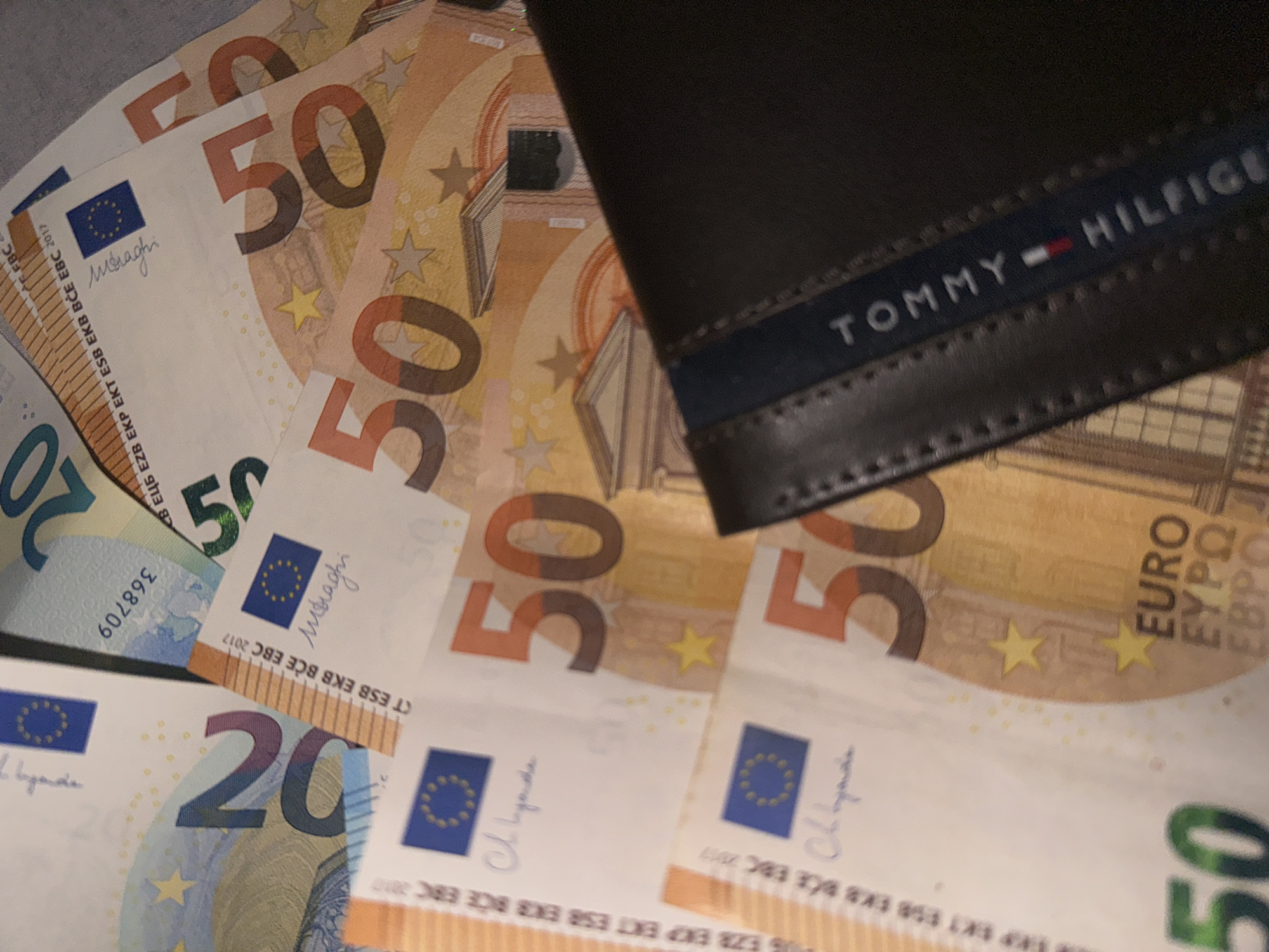 A Tommy Hilfiger wallet is on top of several Euro bills. There are 50 Euro bills and a 20 Euro bill. The bills are spread out, and the wallet is partially on top of the bills.