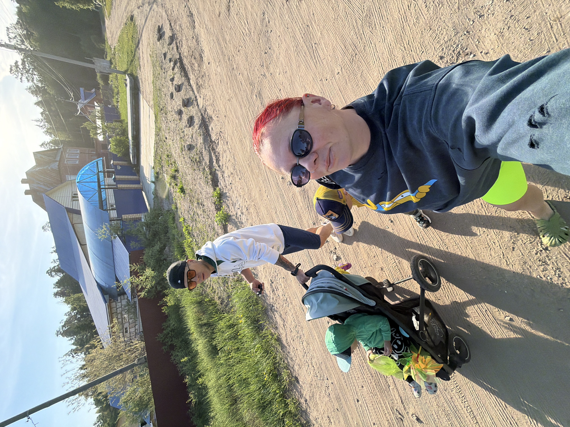 A family consisting of two adults and one child is on a dirt road in Chaikovsky, Russia. One adult is holding the camera and has red hair, sunglasses, and a blue shirt with holes in the sleeve. The other adult is pushing a stroller with a child in it. The adult pushing the stroller is wearing a white sweatshirt, dark shorts, a hat, and orange sunglasses. The child in the stroller is wearing green clothing. The background consists of a fence, grass, and buildings with blue roofing. The scene appears to be captured on a sunny day.