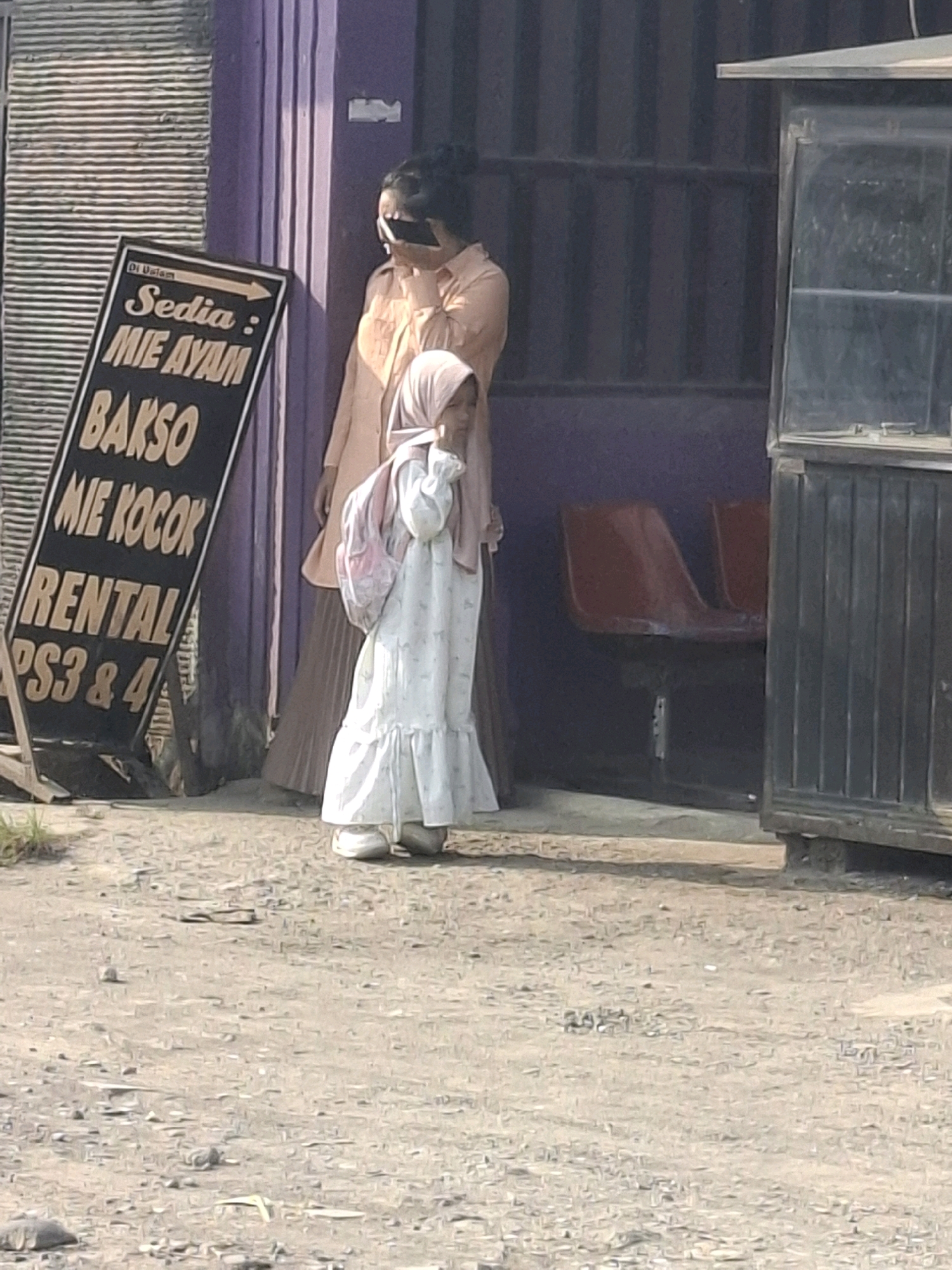 In front of a purple-painted building with a corrugated metal wall to the left and a dark metal structure with a glass display case to the right, a child wearing a white dress and a pink hijab stands on a dirt and gravel ground. The child is carrying a pink backpack and is looking down and to the right. Behind the child, an adult in a light orange shirt and brown skirt is partially visible, holding a dark object to their face. A black A-frame sign is propped up to the left of the child, displaying text in white and yellow font. The sign reads "Di Dalam Sedia: MIE AYAM BAKSO MIE KOCOK RENTAL PS3 & 4". The environment appears to be an outdoor area with some dry grass in the foreground.