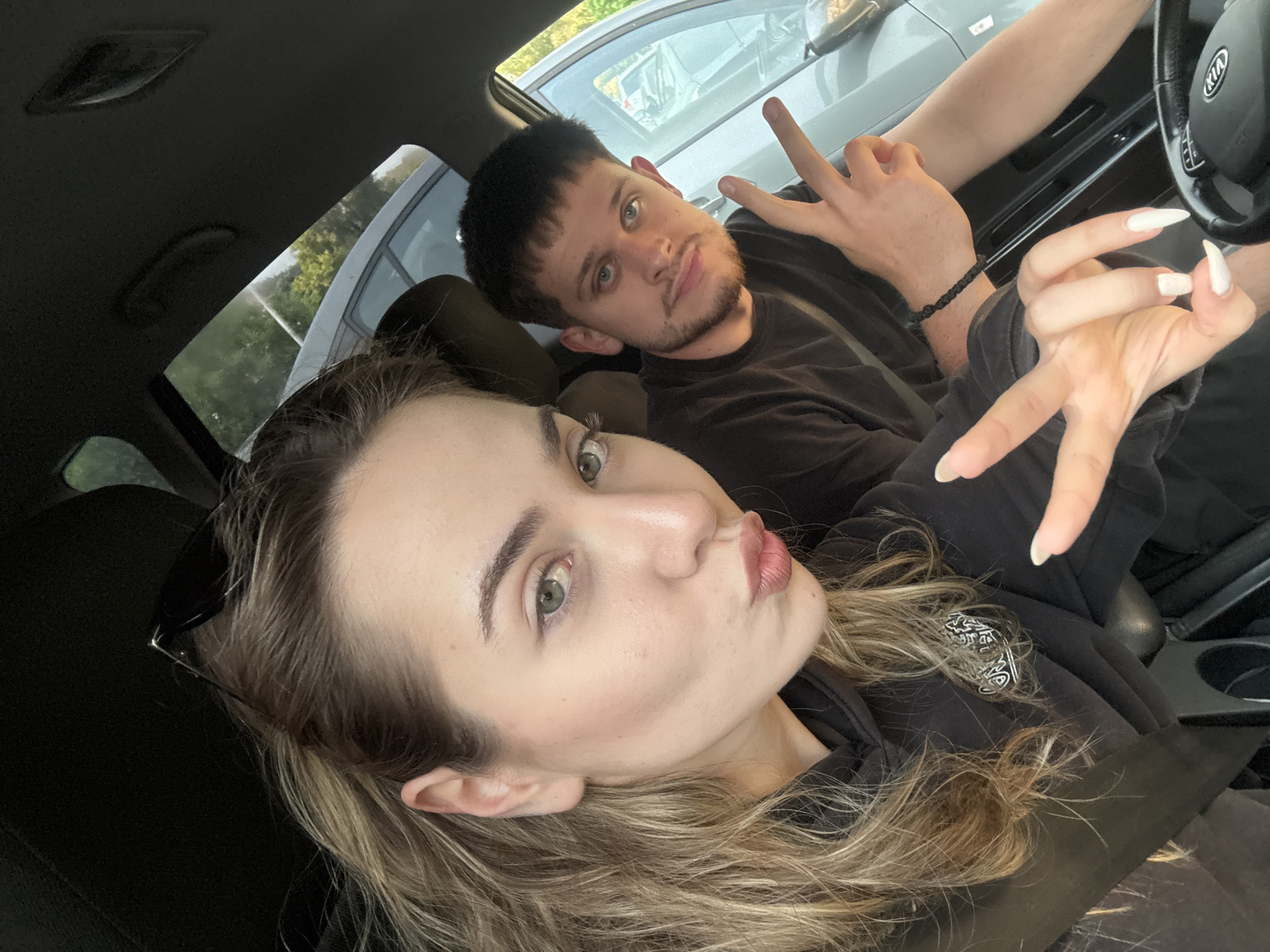 A selfie-style photograph taken inside a car features two individuals. The woman in the foreground, positioned on the left side of the frame, has long, wavy blonde hair, fair skin, and light-colored eyes. She is making a kissing face towards the camera, while flashing the peace sign. Sunglasses are resting on her head. She wears a black hoodie.
The man in the background is to the right, has short, dark hair and facial hair, light-colored eyes, and a neutral expression. He also flashing the peace sign with the right hand. He is wearing a black t-shirt and what appears to be a black bracelet.
The interior of the vehicle, including the steering wheel, Kia logo, and dashboard, is visible. Outside the car windows, another car can be seen.
The location cannot be confirmed as Blagoevgrad, Bulgaria based on the available information.