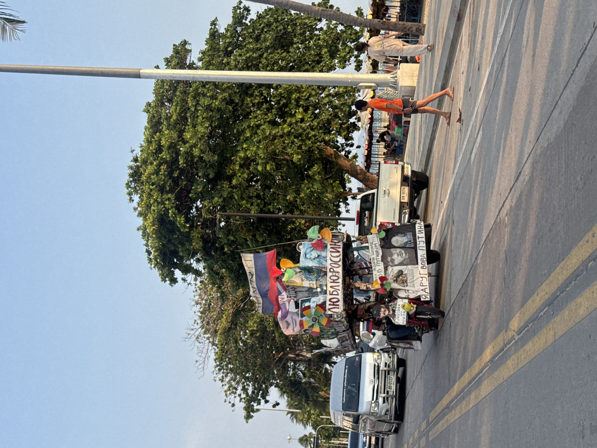The image depicts a street scene in Pattaya City, Thailand. The focus is a vehicle adorned with Russian flags, images of Vladimir Putin, and signage in Russian. The signs contain messages like "I love Russia" and "I am friends with Vova Putin." The vehicle is parked next to a sidewalk, and cars pass on the adjacent road, which is marked with double yellow lines. A large tree provides shade. Pedestrians can be seen on the sidewalk in the background. The sky is clear.