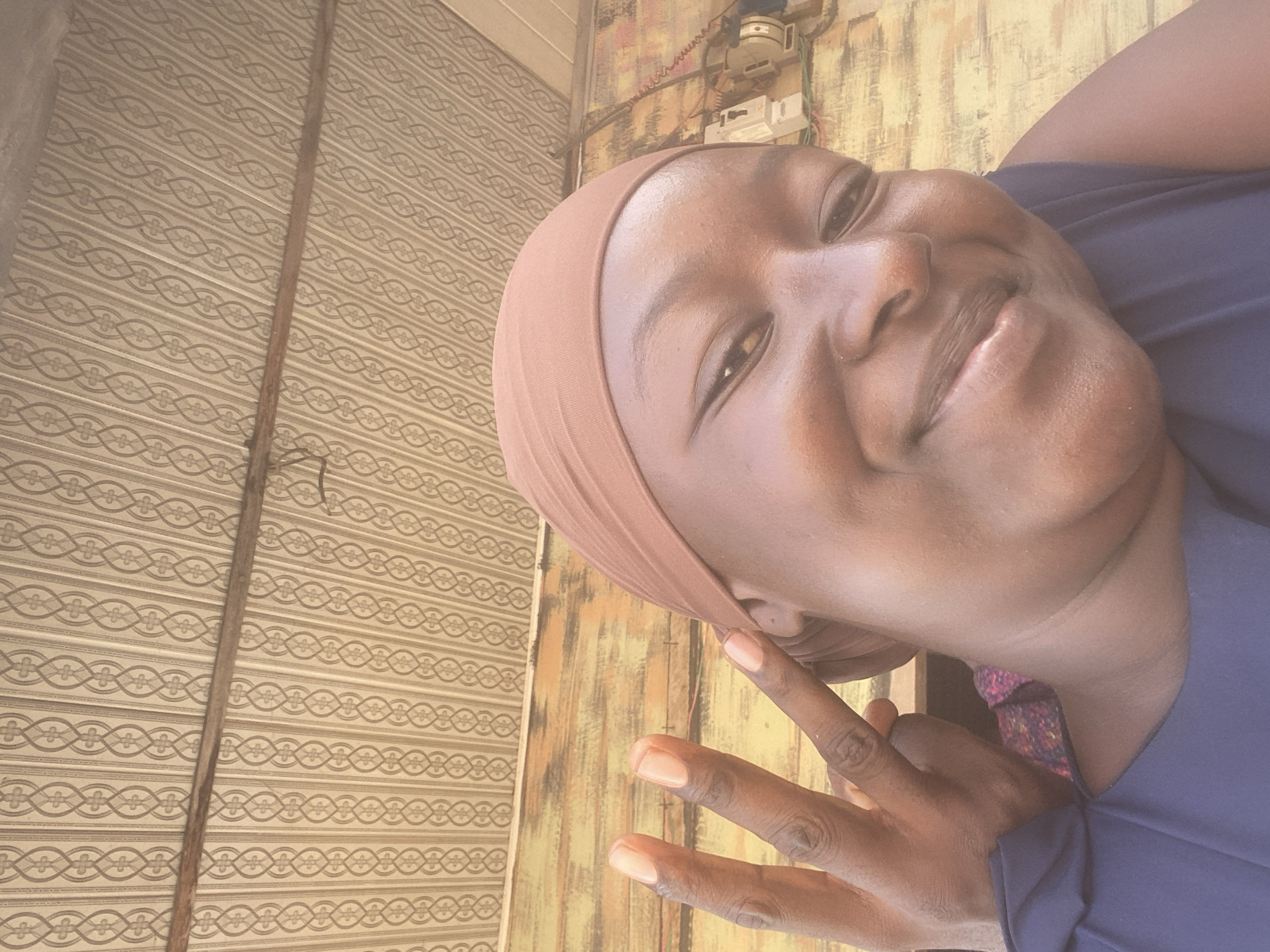 A close-up, eye-level, medium shot captures a Black woman in a selfie. She is wearing a light brown headscarf and a dark blue sleeveless top with a floral pattern visible at the neckline. Her right hand is raised, with her index and middle fingers extended in a peace sign, positioned near her face. Her skin is dark, and she is smiling broadly, with her head tilted slightly. The background consists of a textured ceiling with a repeating pattern and a wooden wall. Behind her head, on the wooden wall, an electrical box with wires and switches is visible. The lighting is natural, suggesting an outdoor or well-lit indoor setting. The scene appears to be from Jalingo, Nigeria, based on the context provided.