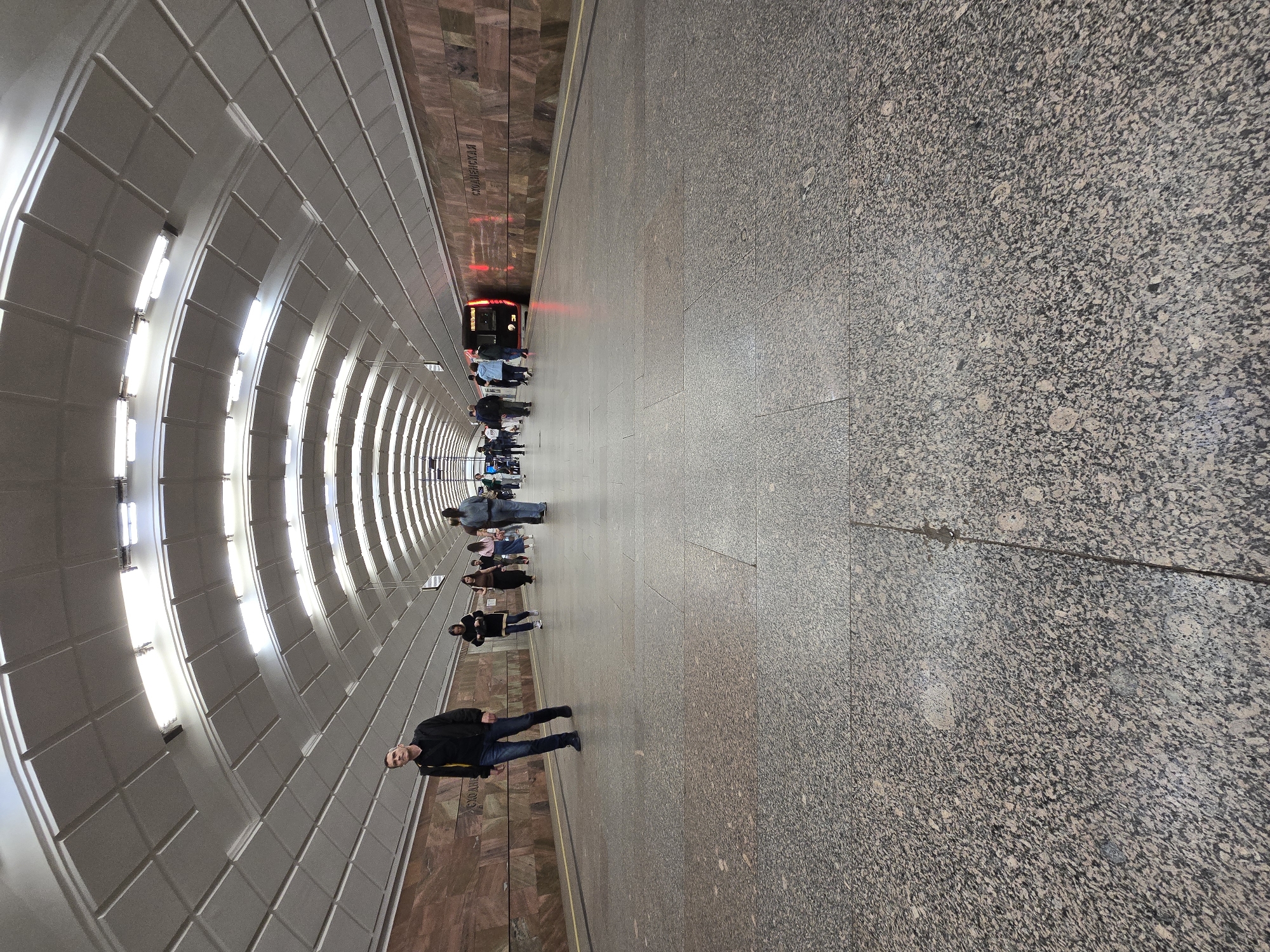 Here's a detailed description of the image:
**Location:** The image appears to be taken in a Moscow Metro station, likely the Smolenskaya station, Russia. The name "СМОЛЕНСКАЯ" is visible on a wall.
**Environment:** It's a large underground space. One side is characterized by a curved, vaulted ceiling composed of repeating white rectangular tiles illuminated by rows of lights. The opposite side has a patterned brown marble wall. The floor is covered with a speckled granite.
**Subjects:**
* Several people are standing on the platform.
* A metro train is visible in the distance with its red taillights illuminated.
* A man wearing a black jacket and blue jeans is standing in the foreground. He is looking at the camera.
**Objects:**
* Metro Train
* Lighting fixtures
* Marble wall
* Granite flooring
* Subway platform
**Actions/Interactions:**
* People are waiting for the train.
* The man in the foreground is standing and looking at the camera.
* The train is stopped at the station.
