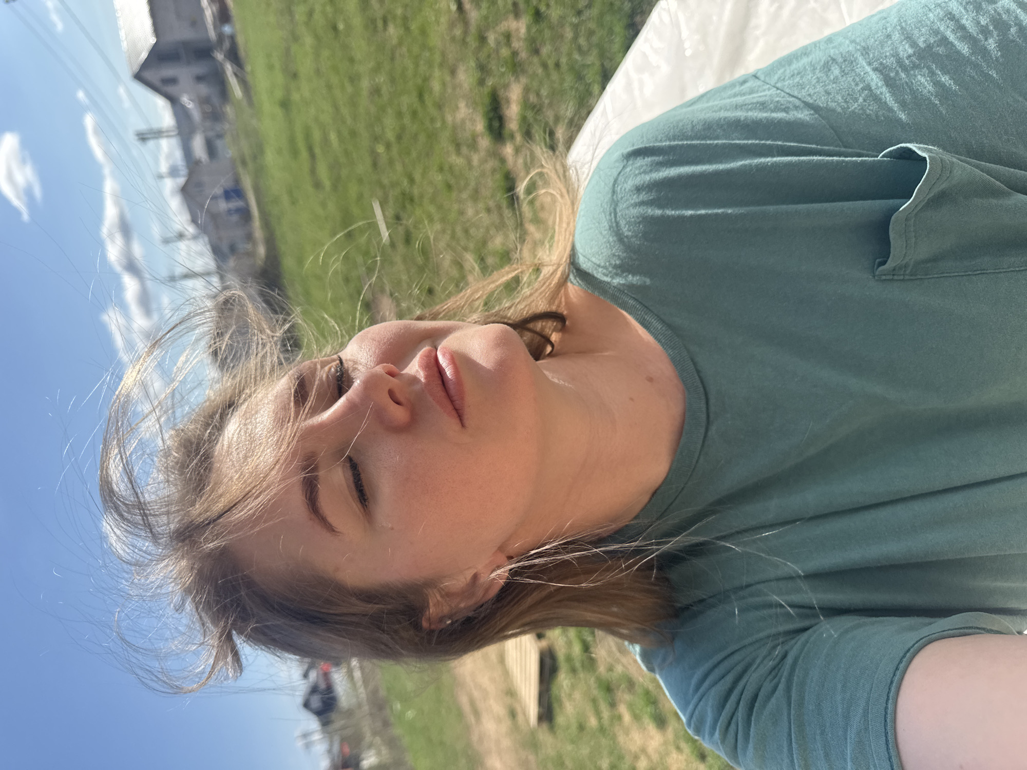 The image is a selfie taken outdoors in Ферма, Russia. A fair-skinned young woman with closed eyes, light brown hair blowing across her face, and wearing a green t-shirt is the primary subject. The background includes a grassy field, a blue sky with some clouds, and a building with power lines in the distance. The camera is tilted slightly. The woman is lying on a white surface.