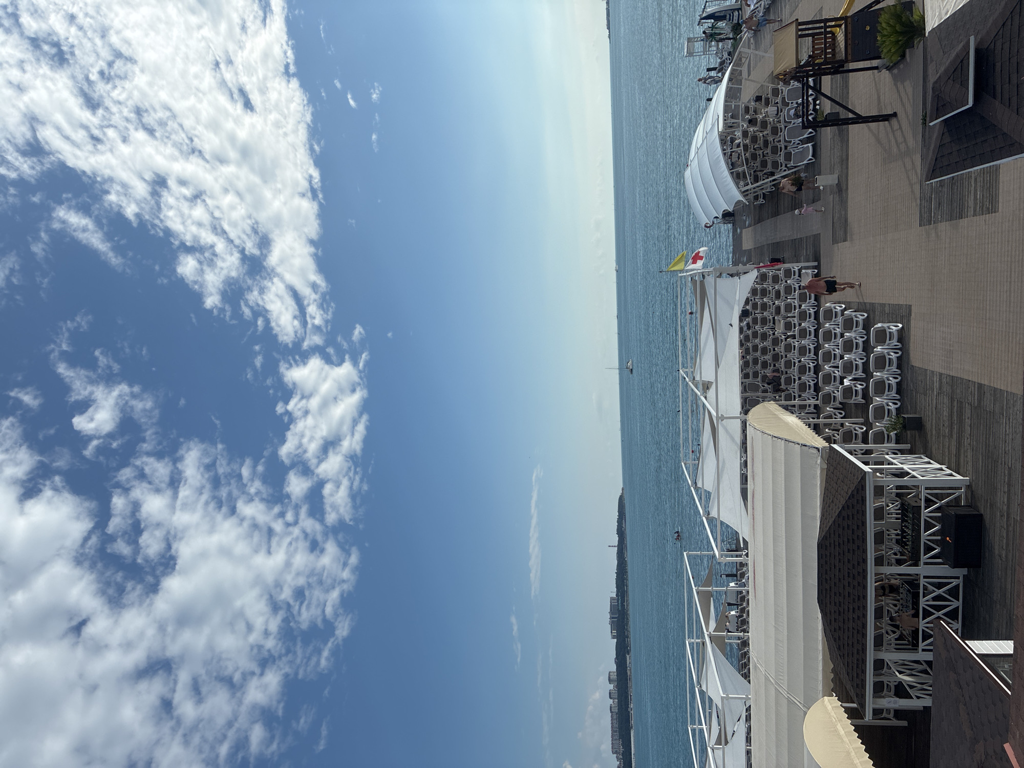 This image depicts a view of the Gelendzhik embankment in Gelendzhik, Russia, on a sunny day with a blue sky and scattered white clouds. In the foreground, there is a wooden pier with rows of empty lounge chairs covered by white canopies. Several people are visible on the pier, including individuals walking and a child with an adult. One white structure with a dark roof and latticework appears to be a gazebo or small cafe, with two people inside. Further back, more canopied lounge chairs are arranged. The turquoise sea stretches to the horizon, where a distant cityscape with buildings and a lighthouse can be seen. A small sailboat is visible on the water. A yellow flag and a white flag with a red cross are flying from a pole on the pier.