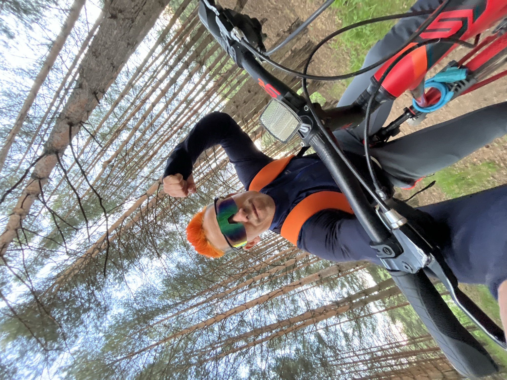 A cyclist, with brightly colored orange hair and reflective sunglasses, poses with a raised fist while riding a mountain bike in a forest in Chaikovsky, Russia. The cyclist wears a navy blue and orange long-sleeved athletic shirt and grey pants. The bike has a red frame with black handlebars and a blue coiled lock attached. The background shows a dense forest with tall, thin trees and a partly cloudy sky.