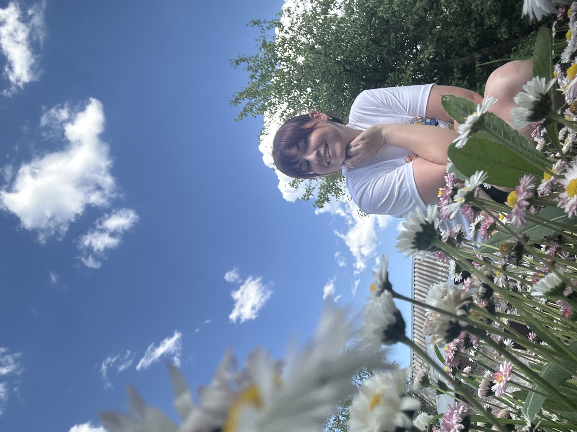 A woman with brown hair and a white t-shirt smiles and looks at the camera. She is surrounded by daisies in front of a clear blue sky with white clouds. A tree is visible in the background. The location is unknown.