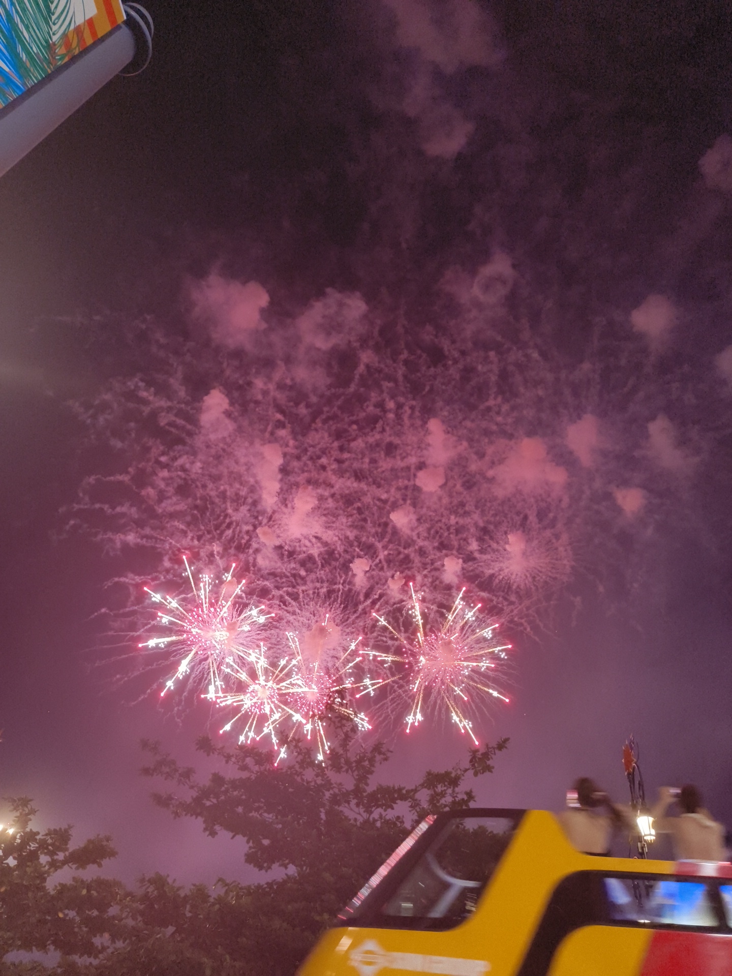 This image captures a nighttime fireworks display over a city, likely Phú Quốc, Vietnam, given the presence of lush foliage. In the foreground, the top of a bright yellow and red vehicle, possibly a tour bus, is visible, with its windows reflecting the colorful lights. Several individuals are seated on the upper deck of the vehicle, silhouetted against the night sky as they observe the fireworks, some appearing to be taking photos. The sky is filled with bright pink and white bursts of fireworks, creating a vibrant and celebratory atmosphere. Wispy pink clouds, a byproduct of the fireworks, are scattered throughout the upper portion of the frame. The lower part of the image shows the dark silhouettes of trees, suggesting an outdoor event.