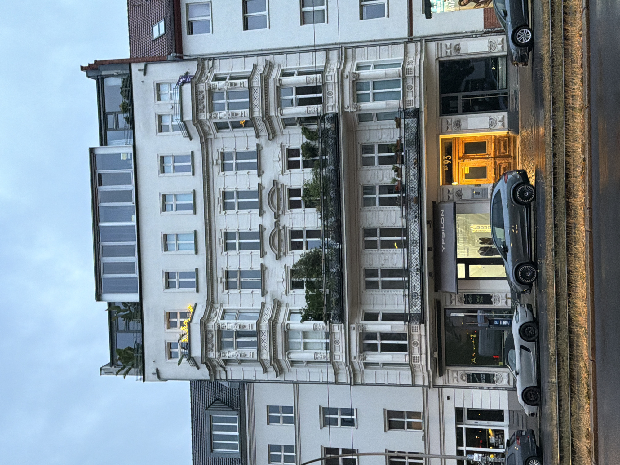 The image shows a white, multi-story building in Berlin, Germany, with a row of cars parked in front. The building features ornate architectural details, including balconies with intricate railings and decorative window frames. The ground floor houses several businesses, one of which has "YPSILON" visible on its awning. A bright yellow door with the number "93" above it stands out. Two cars are visible in the foreground: a light-colored convertible sports car and a dark grey SUV. The sky is overcast, suggesting a cloudy day.