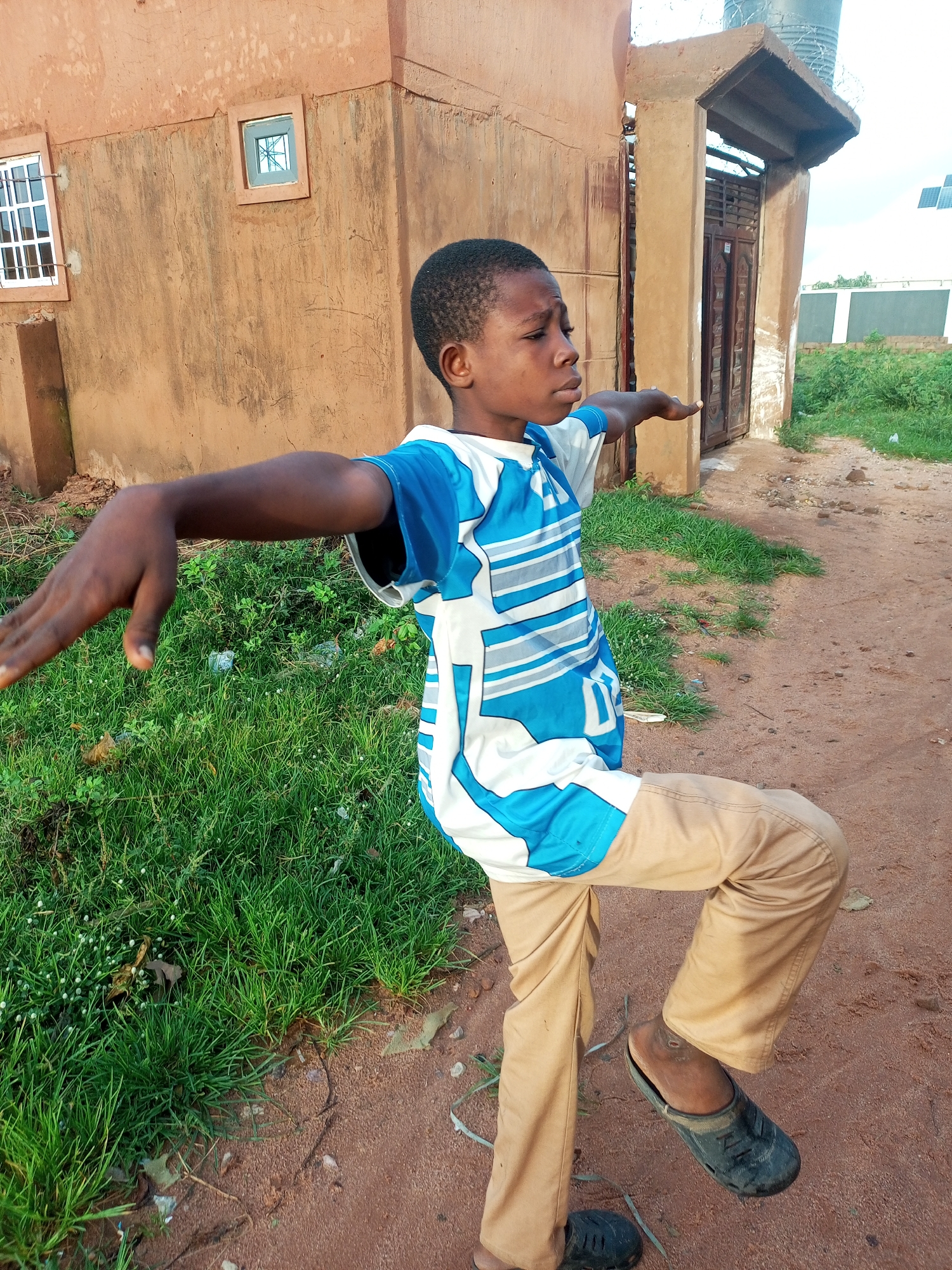 This media file features a young boy in Tempure, Nigeria. He is standing outdoors with a building behind him and grass to his left. The building is made of brown earth-like material with a rough texture. It has a small square window with white bars on the left side and another similar window above and to the right of it. An entrance with a wooden door is visible to the right of the building.
The boy is wearing a blue and white t-shirt with a striped pattern and light brown pants. He is in motion, with his left arm extended to the side and his right leg bent, as if he is dancing or performing an athletic move. His expression is focused.
The ground is made of reddish-brown dirt, with some patches of green grass. The lighting suggests it is daytime, with the sun casting shadows. The overall scene appears to be an outdoor setting in a rural or semi-urban area, consistent with the provided location context. There is no visible text in the image.