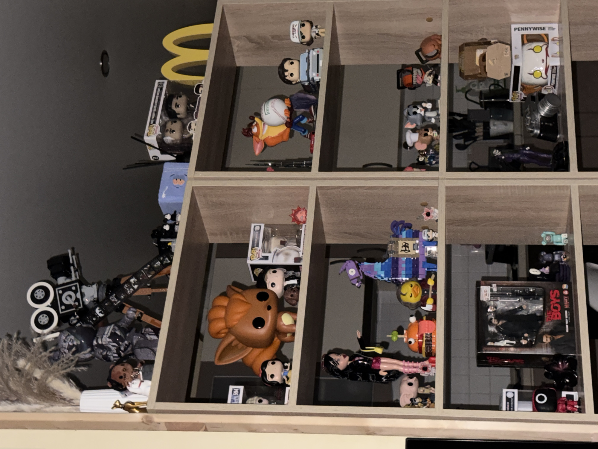 The image shows a wooden shelving unit filled with various collectible figures and items. The shelves are organized into two vertical sections, each with multiple horizontal levels. On the top shelf, in the left section, there is a white vase with dried plants, a small gold figurine, and a plush toy. To the right of these, a large, detailed model of a vintage film camera on a tripod is prominently displayed. Next to the camera, a blue box with a white "B" is visible. The next shelf down, on the left, features several Funko Pop figures, including a large orange creature resembling a fox or dog, a character dressed in black and white, and a figure of Snow White. A small brown dog figure is also present. On the third shelf from the top, left section, a doll with long black hair and a pink outfit stands next to a pig figurine. A hamburger-shaped toy with a face and a yellow duck figurine are also on this shelf, along with a purple llama piñata. The bottom shelf on the left contains a black box with images of people, identified as "THE BOYS," and several smaller Funko Pop figures, including a red figure with a black circle on its head, a black creature, and two small figures on the right. In the right section of the shelving unit, the top shelf displays more Funko Pop figures in boxes, including one with two characters, and a large yellow "M" sign, possibly representing McDonald's. A blue object with a white "B" is also on this shelf. The second shelf from the top, right section, features a figurine of Crash Bandicoot holding a baseball. Next to it are two Funko Pop figures, one in a car and another standing. The third shelf from the top, right section, holds a collection of smaller figurines, including a mouse with a chef's hat, a gray cat-like character, and a figure with an orange chainsaw design. The bottom shelf on the right has a purple figure of the Joker, a black camera, a silver cylindrical object, and a Funko Pop figure of Pennywise. Behind these, there are kitchen utensils in containers, and a cardboard box. The overall environment appears to be an indoor space, likely a room or a display area, with neutral-colored walls. The lighting is dim, creating shadows and highlighting the displayed items. The image is
