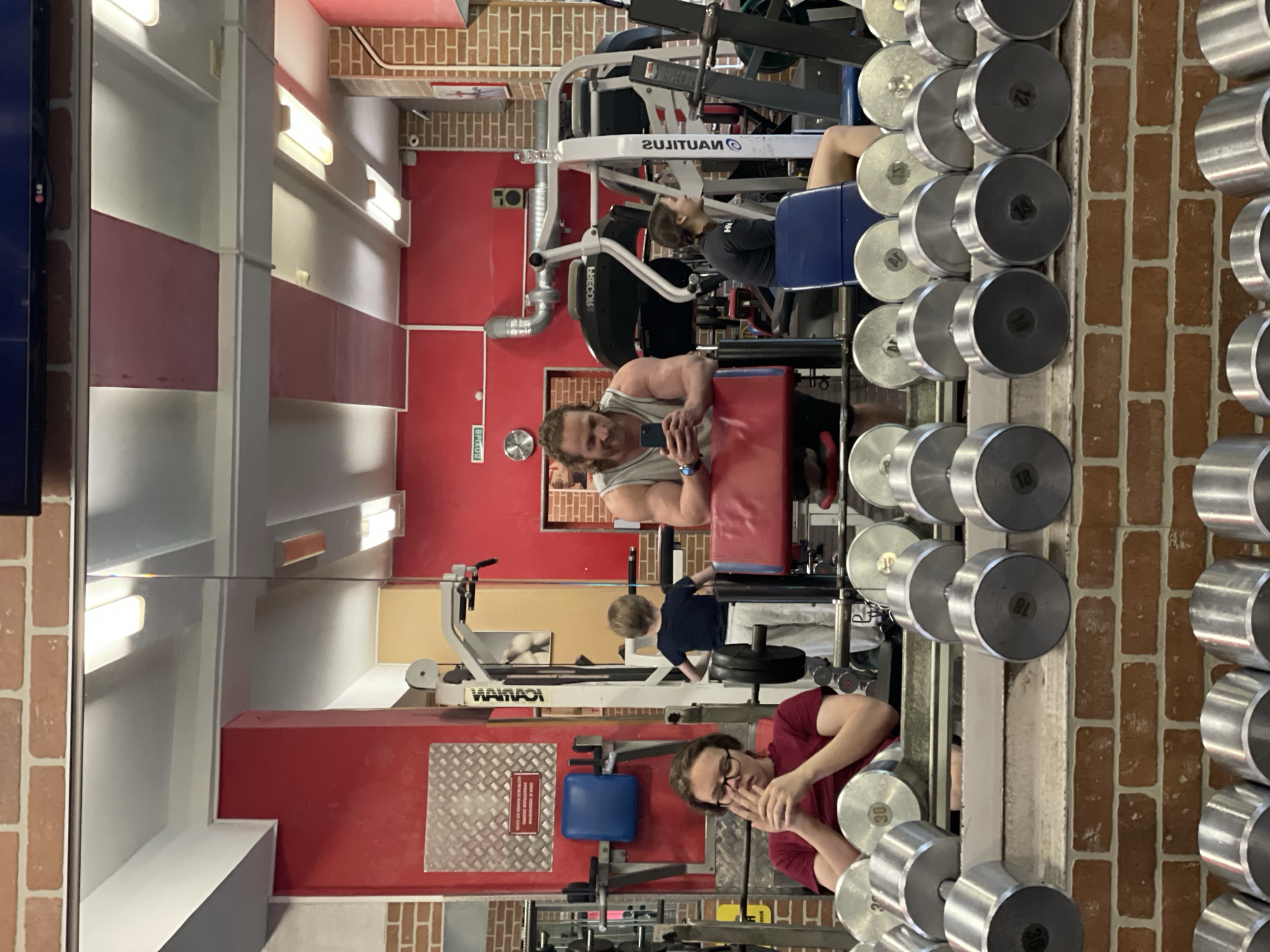 The image depicts the interior of a gym, likely located in Saint Petersburg, Russia, as indicated by Cyrillic text visible on a sign and gym equipment. In the foreground, several rows of dumbbells are neatly arranged on shelves, constructed of metal with visible weight markings. The backdrop is dominated by a large mirror reflecting the gym's interior.
Reflected in the mirror, a man with muscular arms and curly hair is seen holding a smartphone and smiling. He is wearing a grey tank top and appears to be taking a selfie or recording a video. To his right, a woman with dark hair is seated on a blue exercise bench, also reflected in the mirror. A young boy with blonde hair, wearing a dark t-shirt and light-colored pants, stands between gym equipment, facing away from the camera. Another individual, a young person with glasses wearing a dark red t-shirt, is seated in the foreground, also reflected in the mirror, with their hands clasped.
The gym walls are painted in red and brick red, with industrial-style lighting fixtures on the ceiling. Various pieces of gym equipment, including weight machines and benches, are visible throughout the reflected space. The overall environment suggests an active fitness facility.