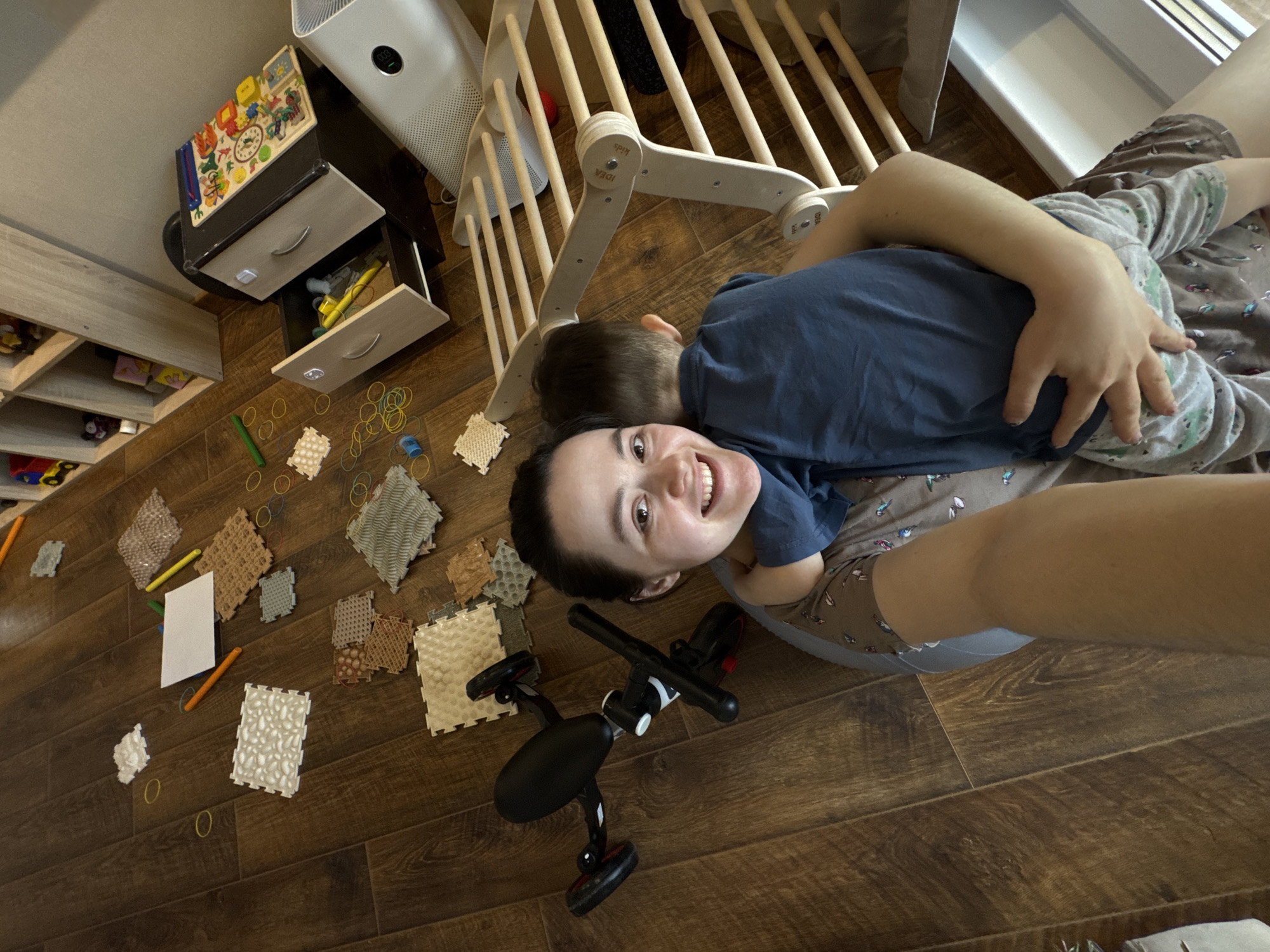 A wide-angle, high-angle shot captures a joyful woman and a young child lying on a dark wooden floor in what appears to be a child's room or play area. The woman, with dark hair pulled back, is smiling broadly at the camera while gently embracing the child, whose head rests on her chest, facing away from the viewer. She is wearing a grey patterned top, and the child is in a blue shirt.
The floor around them is adorned with various play items, including numerous yellow, blue, and green rubber bands, several small textured foam mats of different colors and patterns, a white piece of paper, and orange and green pencils. A black four-wheeled ride-on toy is also visible near the woman.
In the background, to the left, is a light wooden shelving unit with items on its shelves. To the right, a dark wooden dresser with light-colored drawers stands, with some drawers partially open, revealing contents like a yellow pencil or marker. On top of the dresser, a colorful wooden puzzle board with a clock face is placed, next to a modern white air purifier displaying "003" on its screen. A light wooden climbing frame, identified by the text "IDEA Kids" on its structure, is partially visible behind the subjects. The overall scene depicts a warm, playful moment in a home setting, likely in Izhevsk, Russia.