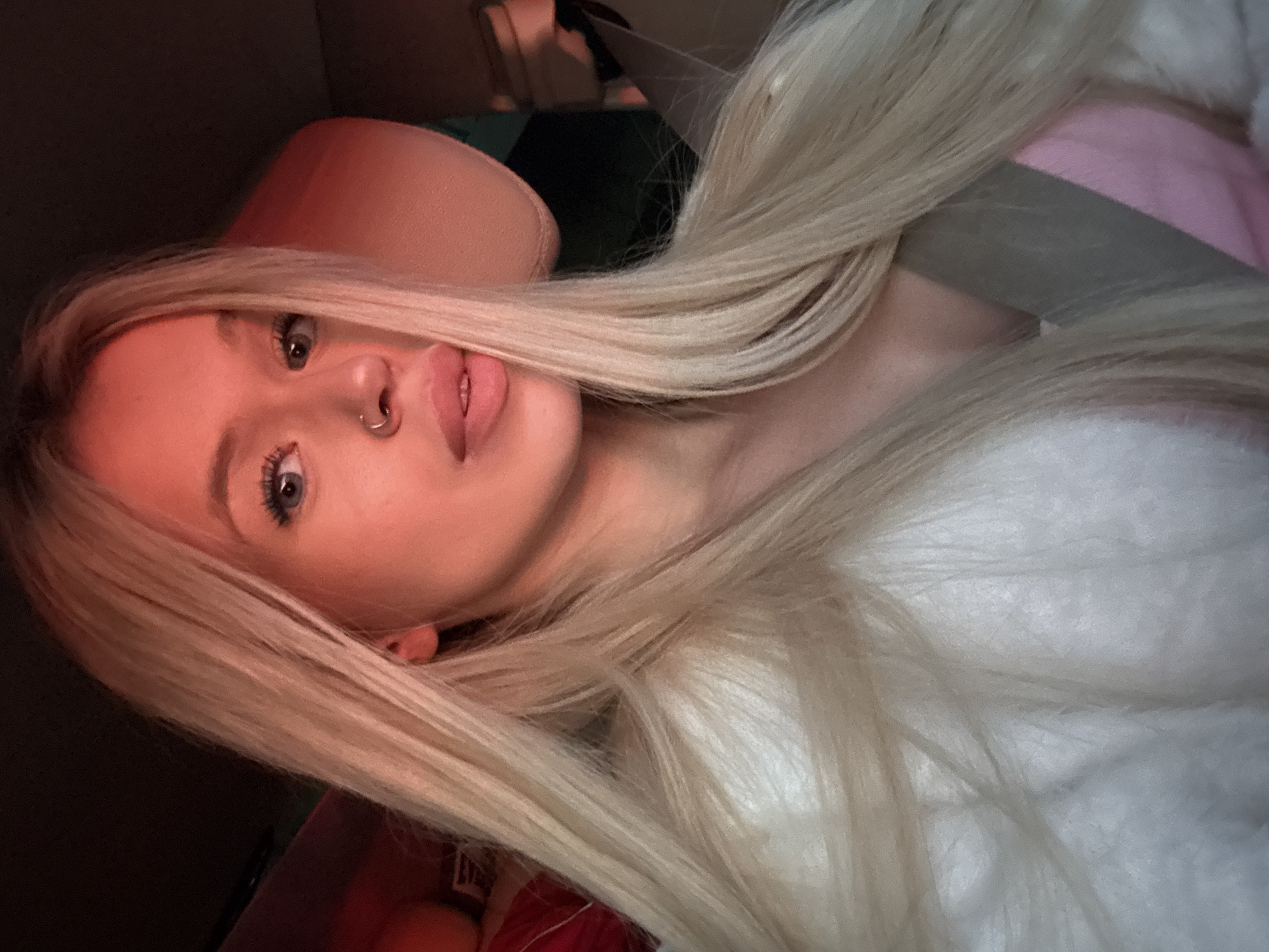 This is a close-up, selfie-style photograph of a young Caucasian woman with long, blonde hair. She is wearing a fluffy white garment over a pink and grey top. She has blue eyes and a nose ring. The lighting in the image is warm and appears to be from an artificial source, casting a pinkish hue on her skin. The background is dark and out of focus, but suggests an indoor environment, possibly a vehicle due to the presence of a headrest. There is no visual information in the image that allows for the determination of a specific location in Saint Petersburg, Russia, or any other geographical area. The image is a portrait and does not depict any actions, interactions, or environmental details beyond what is immediately visible on the subject.