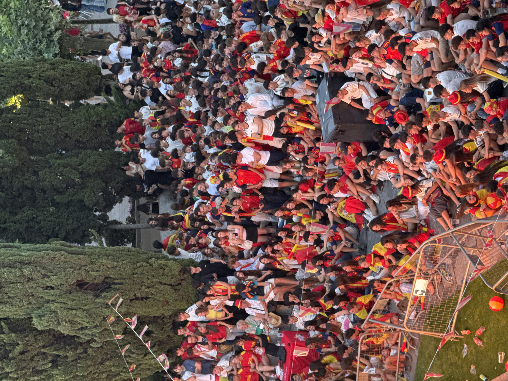 The image depicts a large gathering of people, primarily young adults and teenagers, seated and standing outdoors. Many are wearing red and yellow, the colors of the Spanish flag, and some are adorned with Spanish flags as capes or draped over their shoulders, suggesting a sporting event or celebration. The setting appears to be a public space with mature trees providing shade, and a metal fence encloses part of the area. In the foreground, a patch of green turf is scattered with plastic bottles and small flags. The overall atmosphere seems festive and engaged, with individuals in various states of attention, conversation, and relaxation. The location is identified as the Plaza de España in Las Rozas de Madrid, Spain.