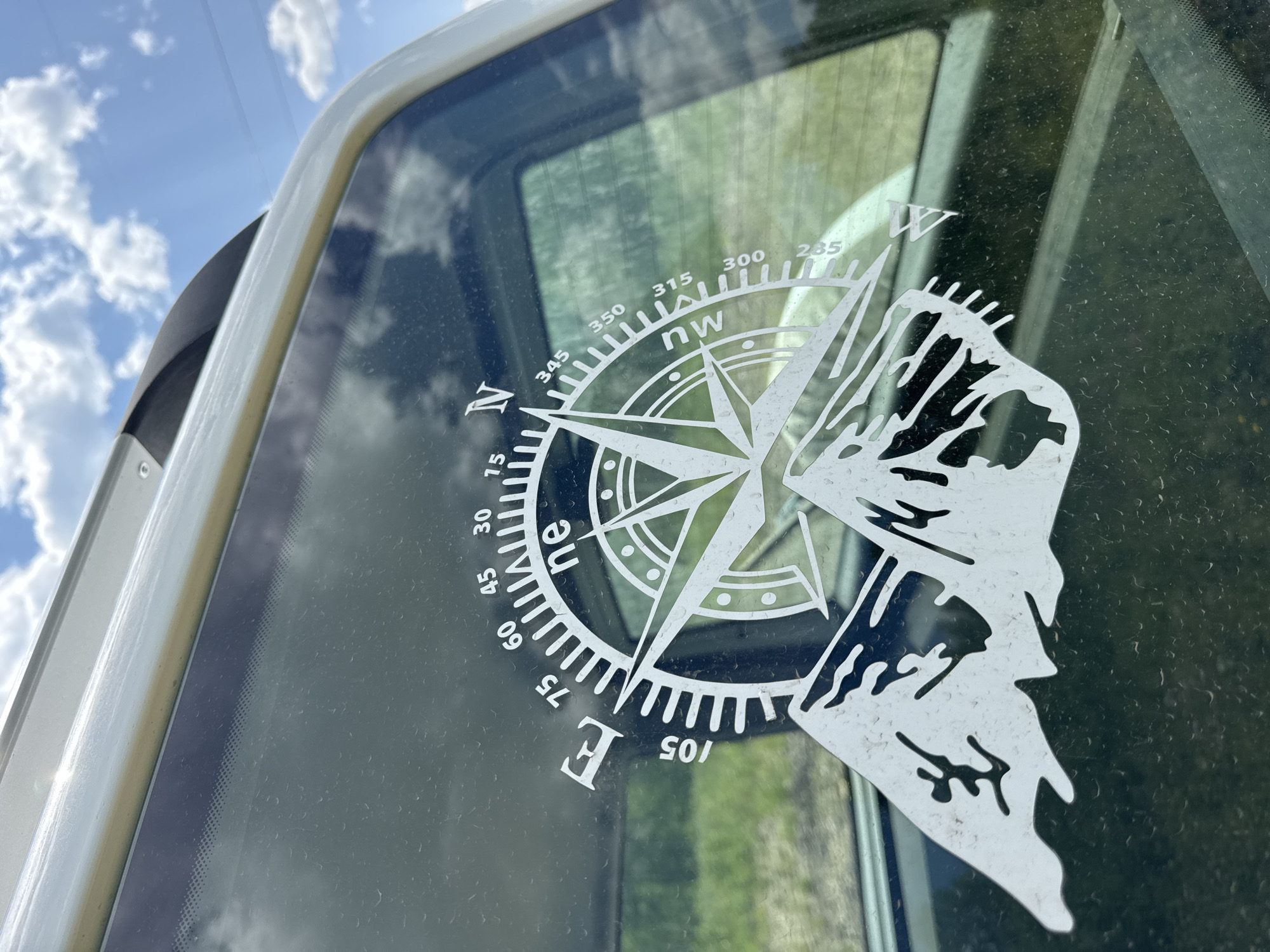 This close-up, angled shot captures a white compass rose decal on a dark, reflective surface, likely a vehicle window. The decal features a detailed compass rose with directional markers and degree increments, along with a stylized mountain silhouette on the right side. The sky, visible in the upper left, is a vibrant blue with scattered white clouds, suggesting a clear, daytime scene. The background behind the decal shows a blurred green landscape, possibly fields or trees. The surface appears somewhat dusty or smudged, with small water droplets visible. The overall impression is one of outdoor adventure and exploration, with the compass and mountain imagery evoking travel and nature. Text visible on the decal includes directional abbreviations such as "N," "NW," and "E," as well as numerical degree markings like "15," "30," "45," "60," "75," "105," "235," "285," "300," "315," and "350." The image is set in Skovorodino, Russia.