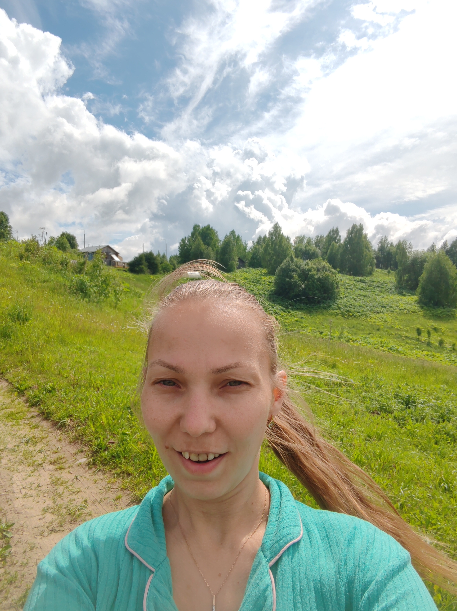 A woman with long blonde hair, wearing a teal top and a necklace, smiles at the camera. She is outdoors in a grassy, hilly area. A dirt path is visible to her left. In the background, there are trees, a house, and a cloudy sky. The scene appears to be in a rural setting.