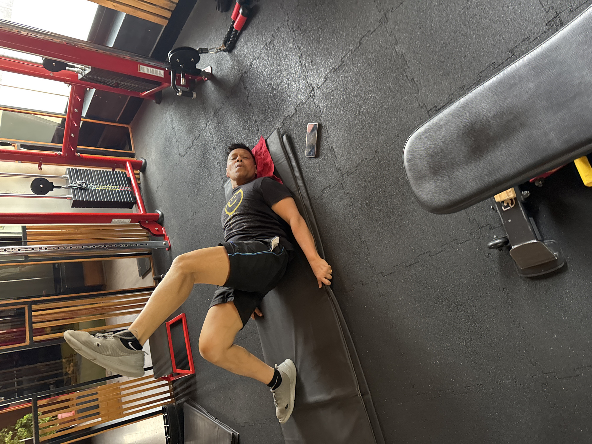 A man is lying on a black mat on a black, rubber-tiled floor in a gym. He is wearing a black t-shirt with a yellow graphic, black shorts with a blue stripe, and grey sneakers with black socks. A red and black weight machine stands to his left, and a black weight bench is to his right. A mobile phone is on the floor near his head. The gym has natural light coming from a window with horizontal blinds on the left side. The location is possibly in Kolkata, India.