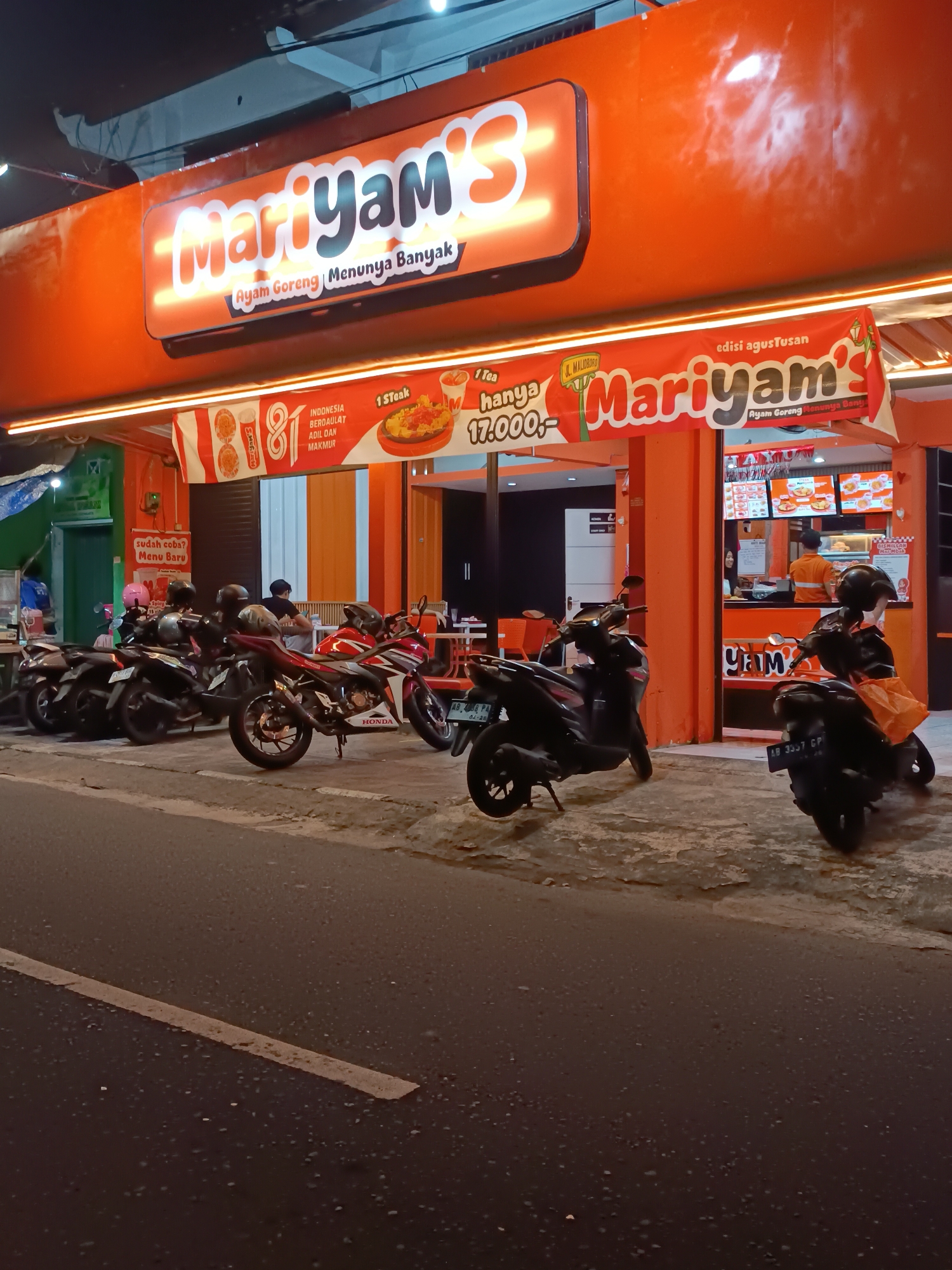 The image depicts a nighttime scene of a "Mariyam's Ayam Goreng" fast-food restaurant, a local chain known for its fried chicken, located on Jalan Malioboro in Yogyakarta, Indonesia. The restaurant's bright orange facade and illuminated signage, featuring the "Mariyam's" logo in neon lights, stand out against the dark sky. A banner above the entrance advertises "edisi agustusan" (August edition) and promotional prices, with an image of fried chicken and fries.
Several motorcycles are parked in front of the restaurant, indicating customer patronage. Some customers are visible inside the restaurant, and at least two staff members are working behind the counter. The street in front of the restaurant is paved, with a white line marking the edge of the road. The overall ambiance suggests a busy evening at a popular eatery in a bustling urban environment.