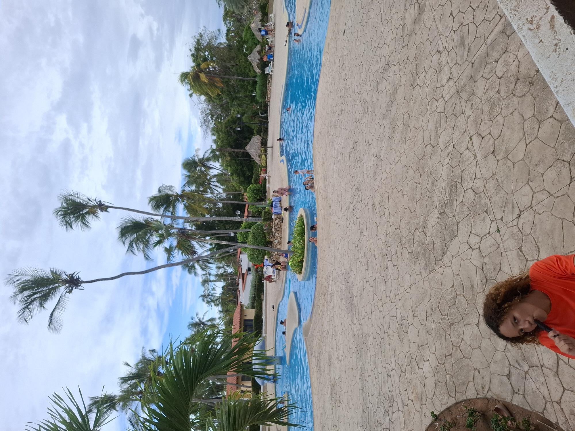 The image shows an outdoor scene at what appears to be a resort or vacation spot, likely in Playa El Agua, Venezuela. A long, winding swimming pool filled with people snakes through the center of the frame. The pool is surrounded by a light tan stone patio. Palm trees are visible along the pool area and extend towards the sky. Some small thatched-roof structures (likely palapas or beach umbrellas) are scattered near the pool. In the lower right corner, a person with curly hair is partially visible wearing an orange shirt. They are holding a dark object to their mouth. The sky is cloudy with patches of blue. The overall impression is that of a relaxed, tropical vacation setting.