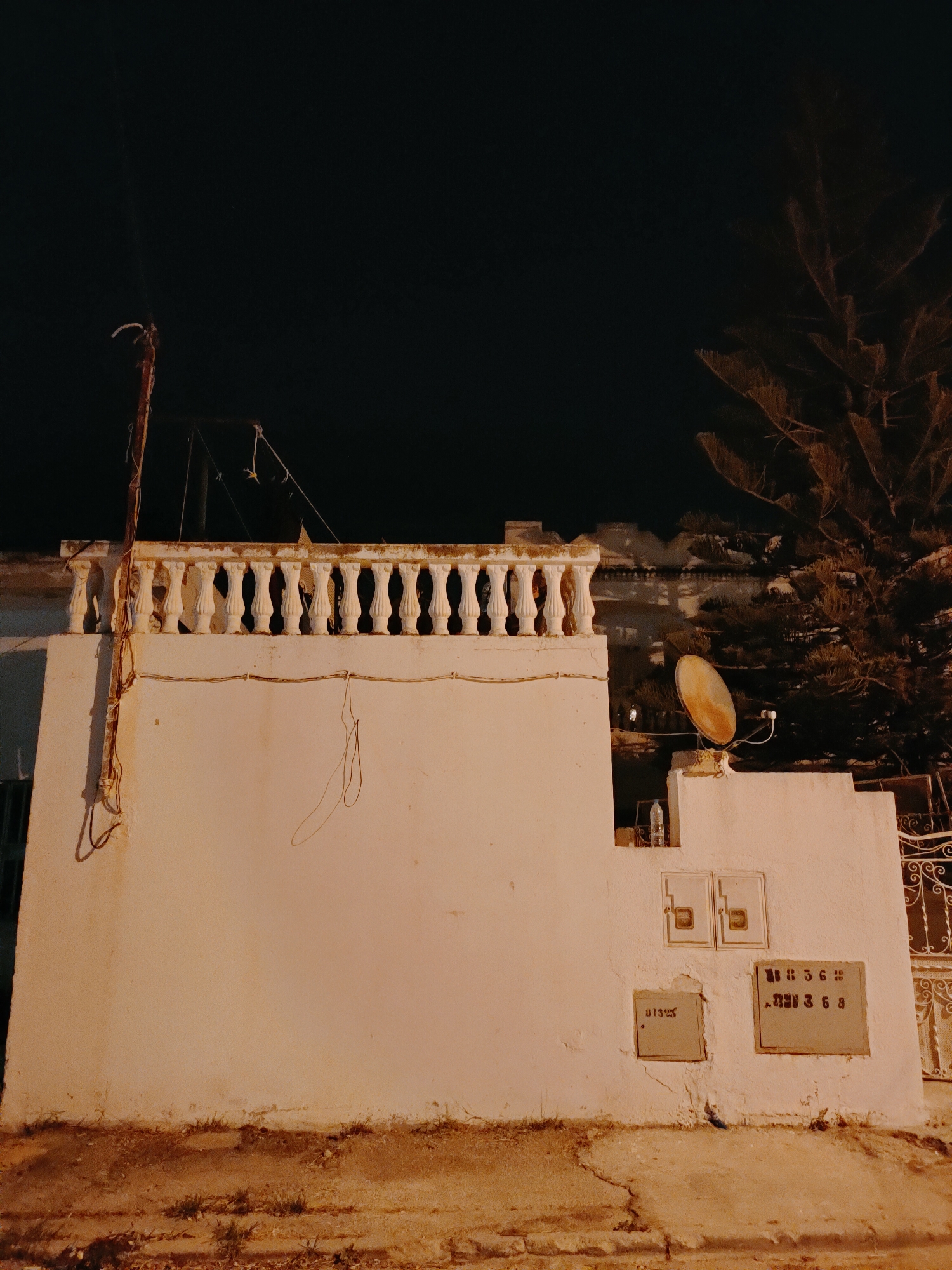 An outdoor night shot captures a low-angle view of a white stucco building's exterior in Tunis, Tunisia. A white ornamental balustrade runs along the top of the building, casting shadows against the dark sky. Below the balustrade, the wall is adorned with several electrical junction boxes and a satellite dish. To the right, a large pine tree with illuminated foliage reaches into the frame. The foreground consists of a rough, dirt ground with sparse vegetation and a cracked concrete pathway. The scene is dimly lit by artificial lights, creating an orange hue across the surfaces.