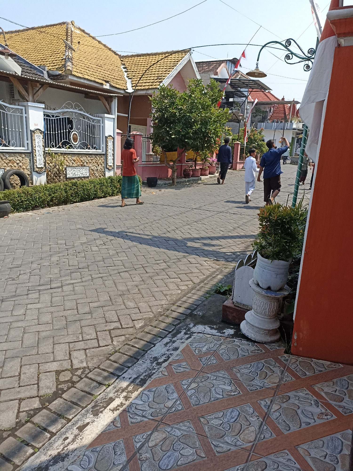 The image shows a street scene in the city of Sumberejo, Indonesia. The street is paved with grey interlocking pavers and lined with residential buildings and greenery. The buildings have tiled roofs and decorative fences. Several people are visible walking on the street. One person in the foreground is wearing a red shirt and a green sarong, walking away from the camera. Further down the street, three other individuals are walking towards the right of the frame. Two of them appear to be young males, one wearing a white outfit and the other a dark blue shirt. The third person is wearing a dark blue long-sleeved shirt and dark pants. Indonesian flags are visible on some of the buildings, suggesting a celebration or national event. The sky is clear and blue, indicating a sunny day. The overall scene depicts a residential neighborhood in a tropical climate.