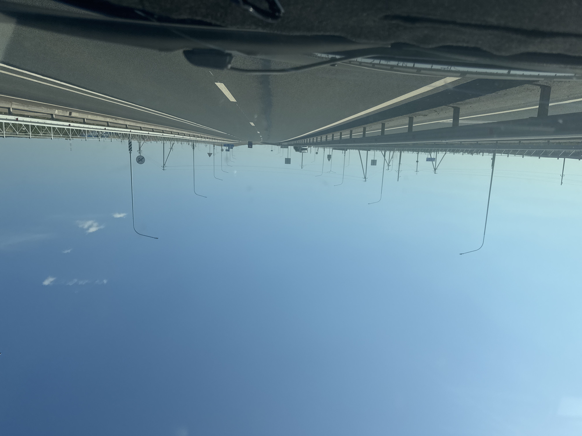 The image is a first-person perspective from inside a vehicle, looking out at a multi-lane highway under a clear blue sky. The road appears to be a modern highway with concrete barriers and metal guardrails along the sides. There are numerous streetlights lining the highway at regular intervals. In the distance, there are some utility poles with power lines, and a few vehicles are visible on the road, including what appears to be a truck. The landscape beyond the highway is flat and appears to be rural or undeveloped. The overall impression is one of a typical highway scene on a bright, sunny day. The image does not contain any specific landmarks or clues that would indicate its location within the city of Syrt, Russia.