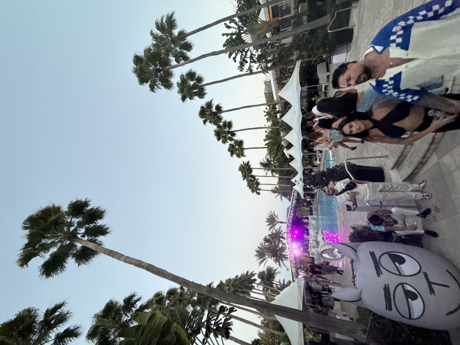 The image displays an outdoor event at a resort pool area in Miami Beach, United States of America, observed during dusk or early evening. The sky is clear and light blue, transitioning to a lighter shade near the horizon.
A large swimming pool occupies the center-midground, reflecting purple light. At the far end of the pool, a stage or performance area is visible, illuminated with bright pink and purple lights and decorated with green foliage. Numerous tall palm trees with green fronds are prominent throughout the scene, framing the area and extending into the background.
Multiple white canopy tents are erected around the pool deck, under which groups of people are standing and mingling. A multi-story building, likely part of the resort, with white balconies or awnings, is visible in the background on the right. Paved walkways and steps are present around the pool.
In the immediate foreground, on the left, a large, grey inflatable sculpture depicting a cartoonish animal head with large eyes and a simple mouth is positioned. A man in a white t-shirt and white pants, wearing a black fanny pack, stands beside a white waste receptacle near the center foreground. To his left, another man in white pants and a patterned shirt is seated on a low barrier or step, looking downwards.
On the right side of the foreground, a woman with dark hair in a black halter-neck top and dark bottoms stands next to a man with a dark beard, who is wearing an open blue and white checkered shirt over a lighter garment. Both individuals are partially turned away from the camera's direct view.
Throughout the scene, approximately two dozen or more individuals are visible, standing, walking, and conversing around the pool, under the tents, and near the stage. A white electric fan unit is positioned near the pool edge on the left, behind the inflatable sculpture.