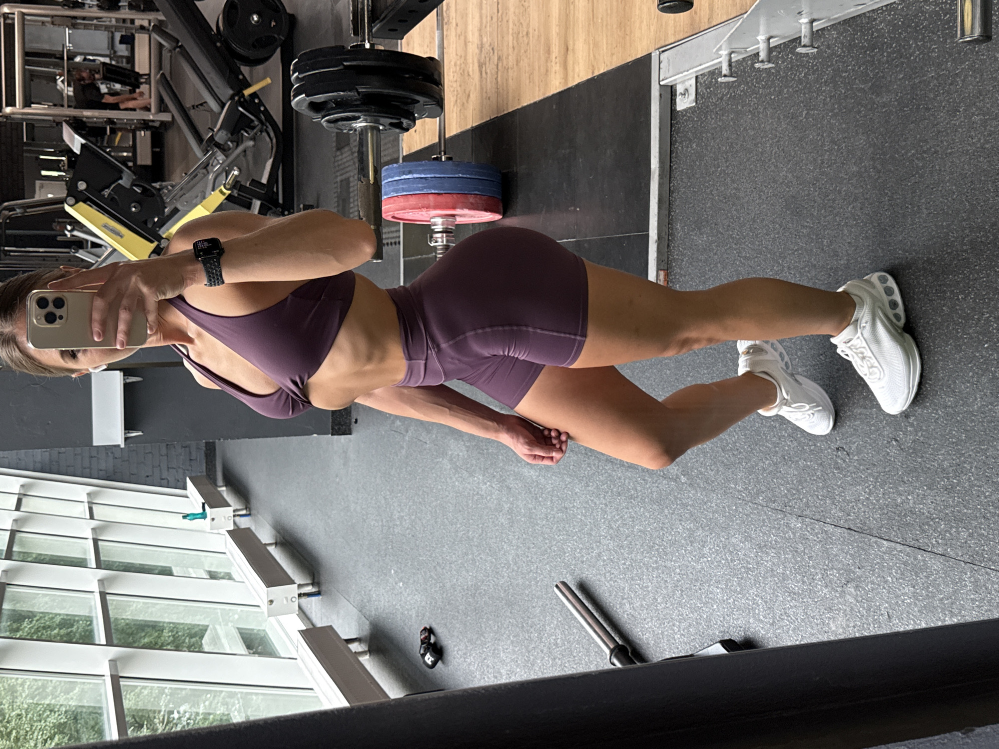 A woman is standing in a gym, taking a selfie with her phone. She is wearing a purple sports bra and shorts, and white sneakers. She has a watch on her left wrist. In the background, there are various gym equipment, including weight machines, barbells, and weight plates. Large windows with a view of trees are on the left side of the image. The floor is made of gray rubber mats, and there are wooden platforms for weightlifting. The overall environment suggests an indoor fitness facility. The image does not contain any specific information that can pinpoint its location to Saint Petersburg, Russia.