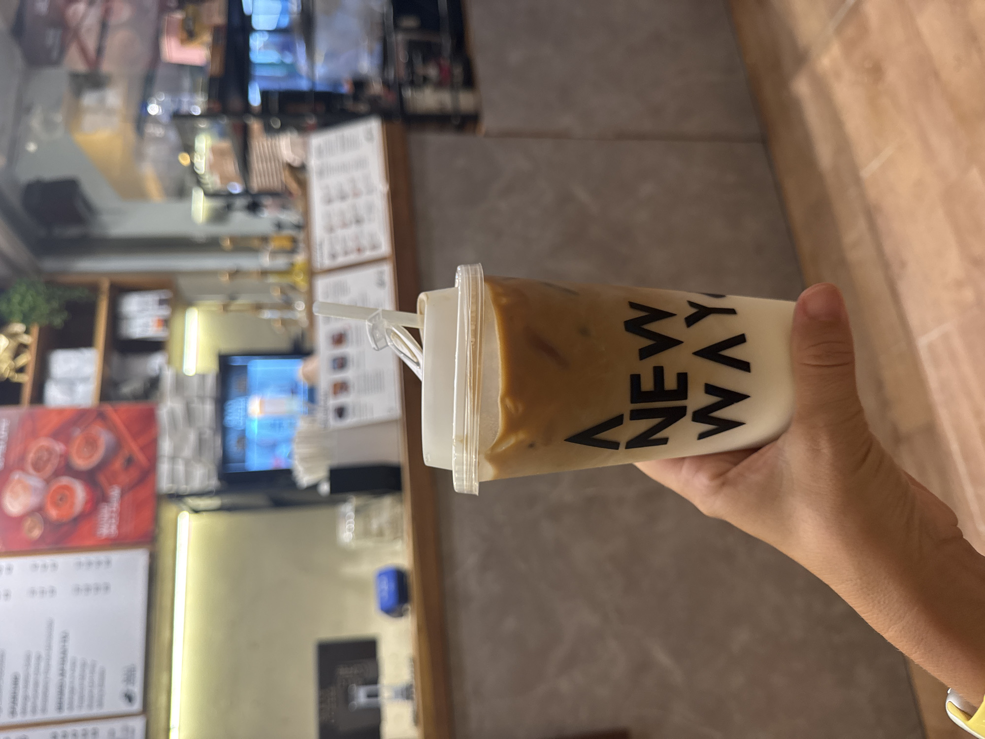 A person holds a plastic cup with a brown and white liquid, labeled "NEW WAY", in front of what appears to be a coffee shop in Kuta Selatan, Indonesia. The shop has a menu board on the left with food and drink photos. A screen behind the counter is displaying information, and additional menus are visible on the right. The environment appears to be inside the coffee shop. The person's hand and part of their arm are visible. They are wearing a yellow wristband.