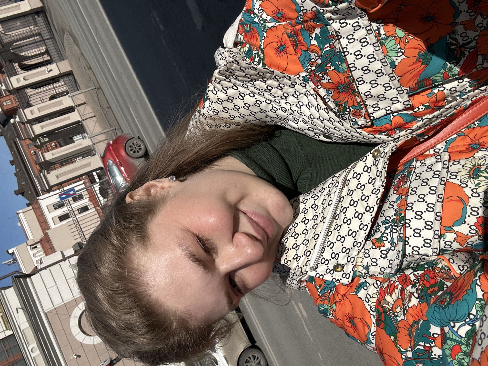 A medium shot depicts a woman outdoors in Kazan, Russia, with a partial view of city buildings in the background. The woman's head and shoulders are the primary focus. She is wearing a green turtleneck sweater covered by a floral-patterned jacket with orange and turquoise accents over a white base, patterned with a repeating black “G” logo. The jacket's zipper is visible. The buildings in the background are multi-story, with architectural details and windows, and the sky above is blue. A red car is visible on the street.