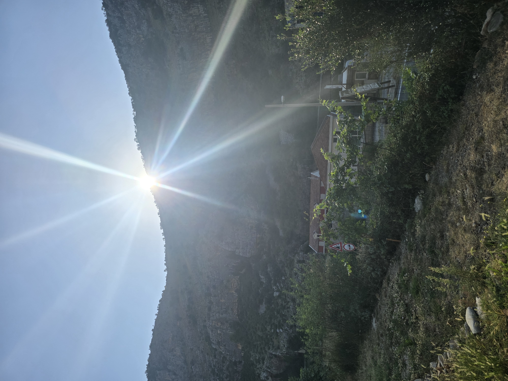 The image depicts a sun flare obscuring the sun behind a mountain, with a town nestled at its base. The sun's rays emanate intensely across the sky. In the foreground, lush green foliage and dry grass cover the terrain. A few houses with red roofs are visible in the mid-ground, partially obscured by trees. A traffic sign showing a truck and a pedestrian is also present. A lamppost stands near the houses. The background is dominated by a large, rocky mountain, with sparse vegetation clinging to its slopes. The overall scene suggests a rural or semi-rural setting outside of a city. The image is likely taken in the vicinity of Podgorica, Montenegro, given the geological features and the architectural style of the houses.