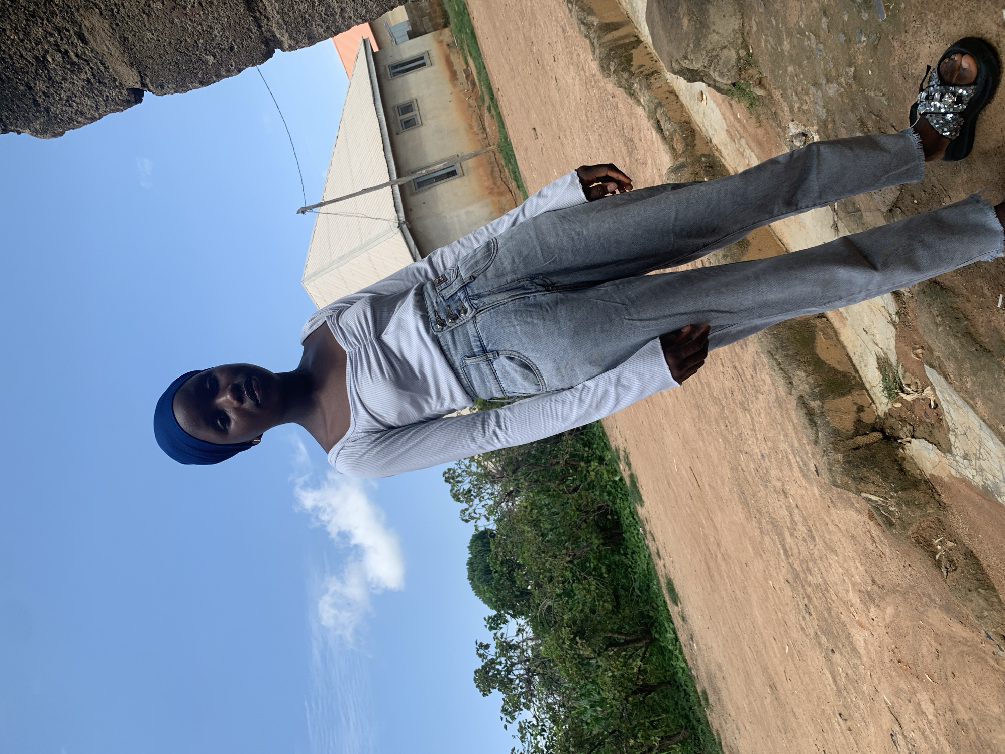 A young woman wearing a blue headscarf, a light grey long-sleeved ruched top, and light blue high-waisted jeans stands in an outdoor environment. She is also wearing black sandals with embellished straps. The background includes a dirt path, overgrown green vegetation, a building with a light-colored roof and brown walls, and a clear blue sky with a few wispy clouds. A rough, stone-like structure is visible to the right of the frame. The scene appears to be a residential area in Sabon Tasha, Nigeria, given the architectural style of the building and the general landscape.